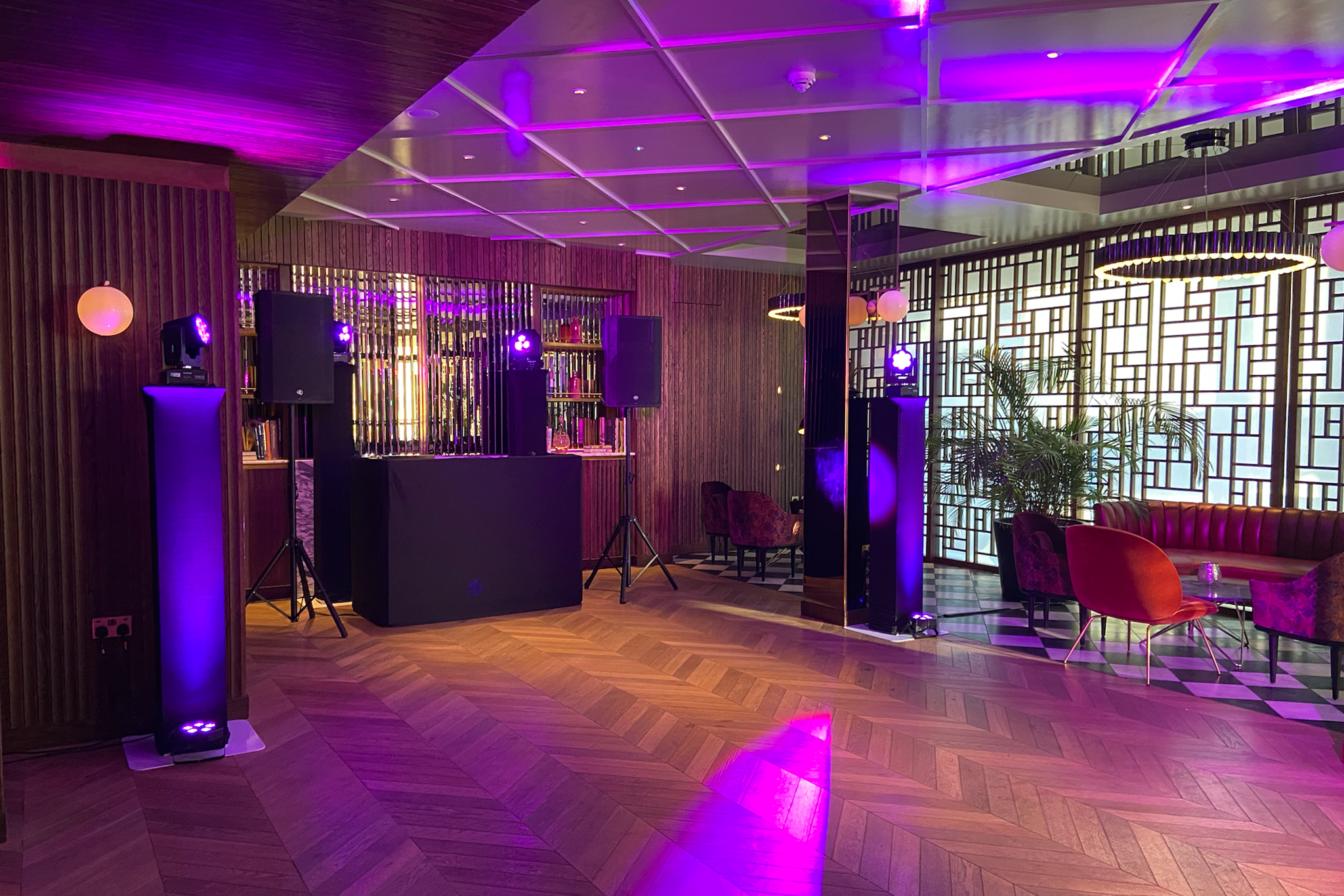 Interior of a modern lounge or event space with purple lighting, wooden panel walls, patterned carpet, and seating areas with chairs and a sofa, some with small tables and decorative items.