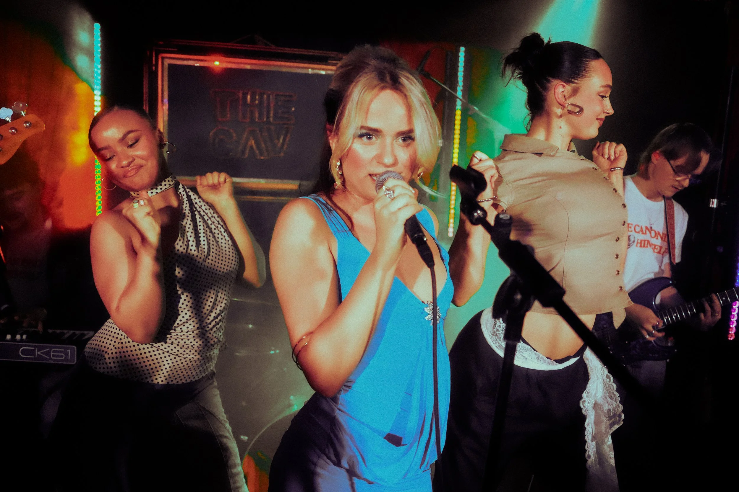 A group of four women performing on a stage with musical instruments and colorful lighting. One woman at the center is singing into a microphone, another is playing guitar, and the others are engaged in the performance.