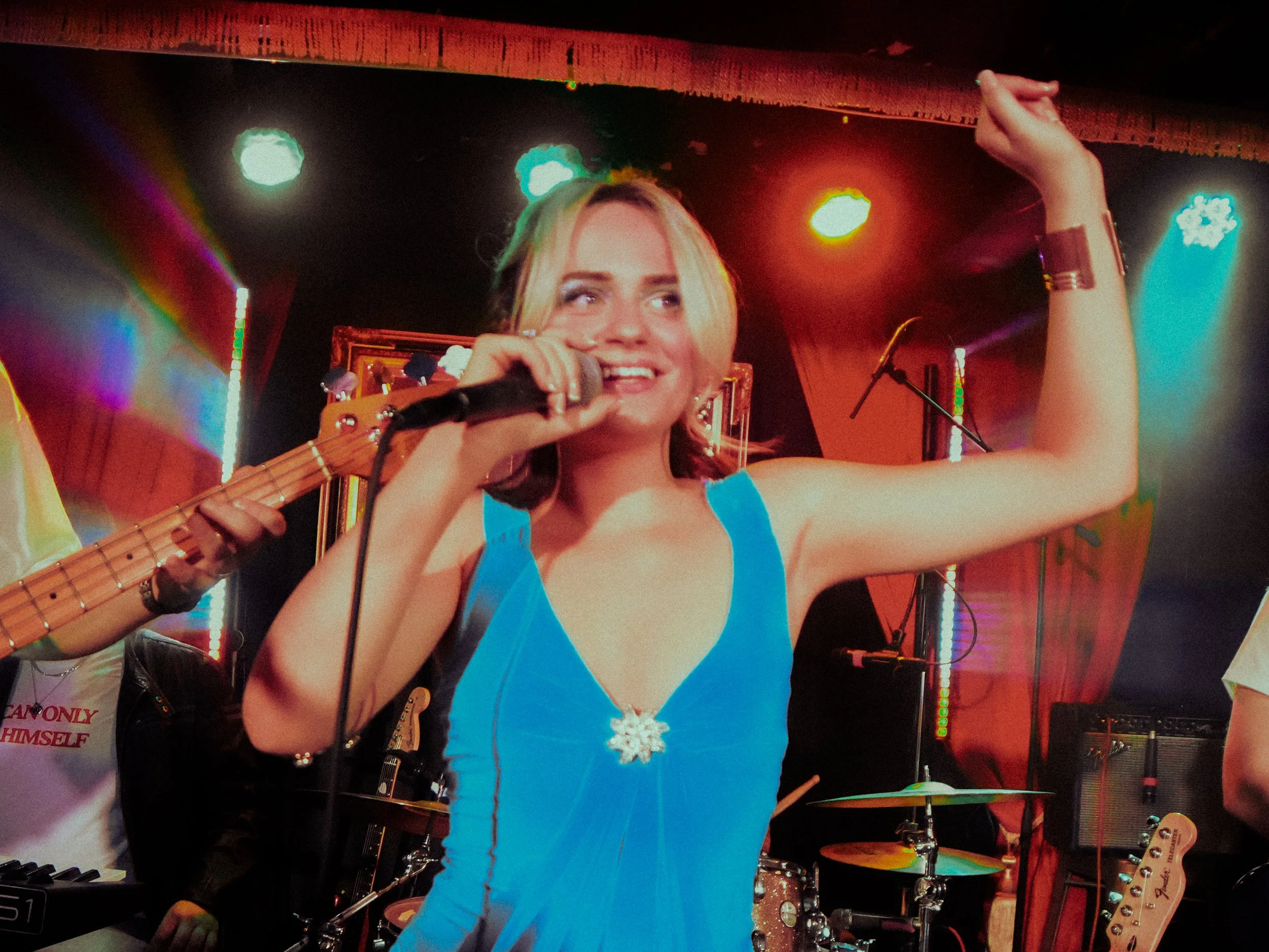 A young woman with blonde hair wearing a blue dress with a snowflake necklace, singing into a microphone on stage. There are colorful stage lights and musical instruments in the background.