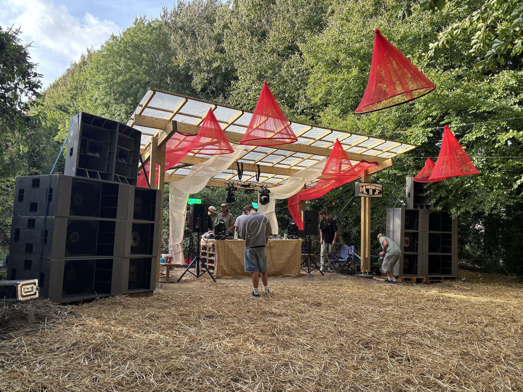 Outdoor music stage at a festival with sound equipment, red cone-shaped decorations, and several people setting up or preparing for a performance surrounded by trees.