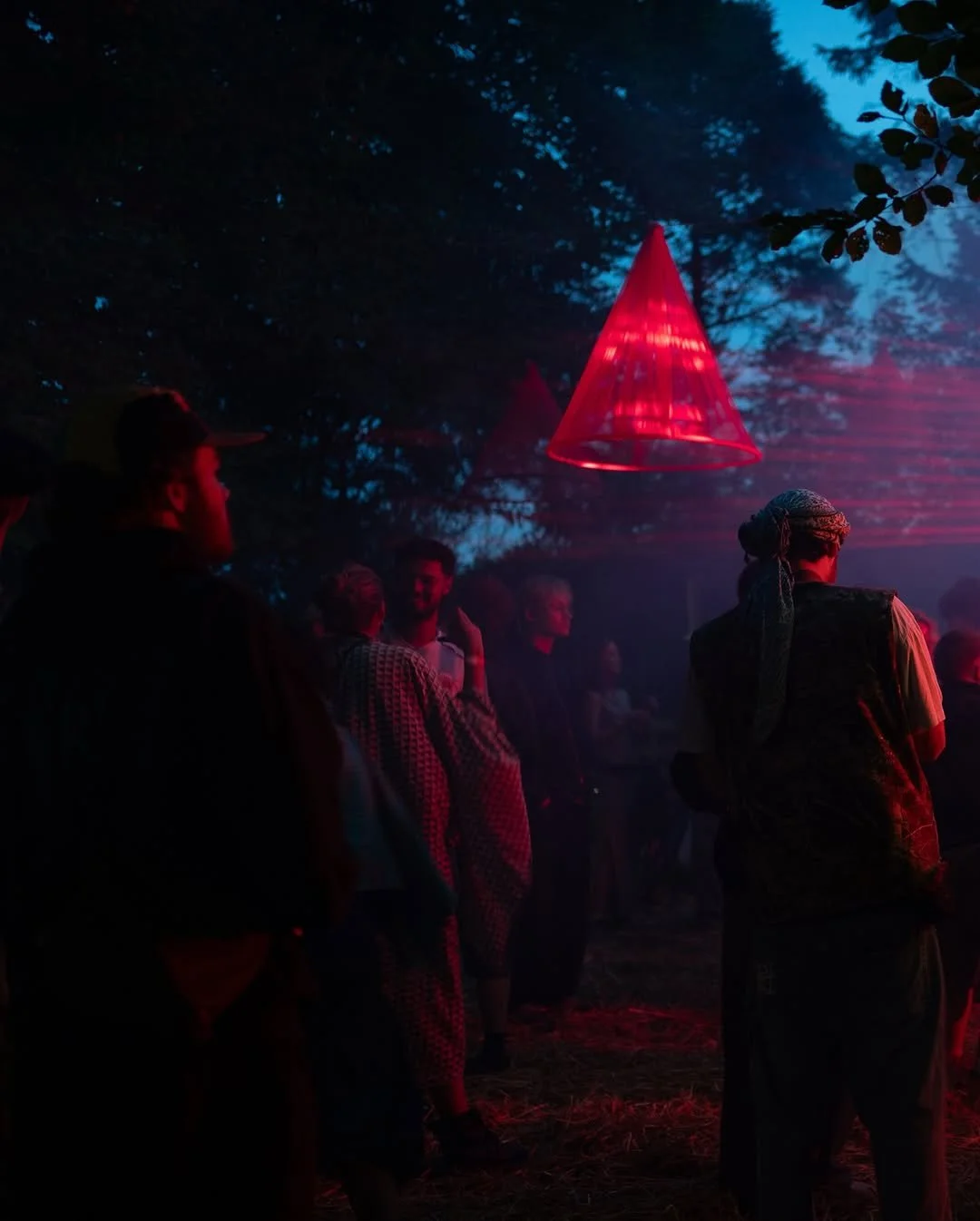 People gather outdoors at night under a red cone-shaped light fixture, with trees and a dark blue sky in the background.