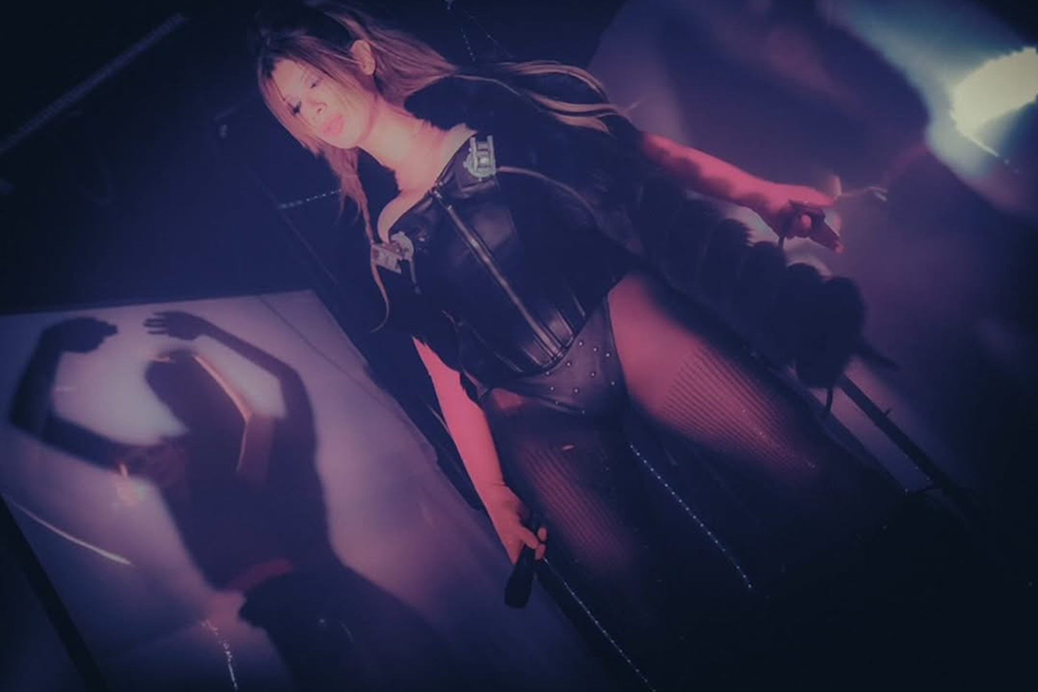 A woman with long hair in a black leather jacket, standing next to a motorcycle in a dimly lit environment with purple and pink lighting.
