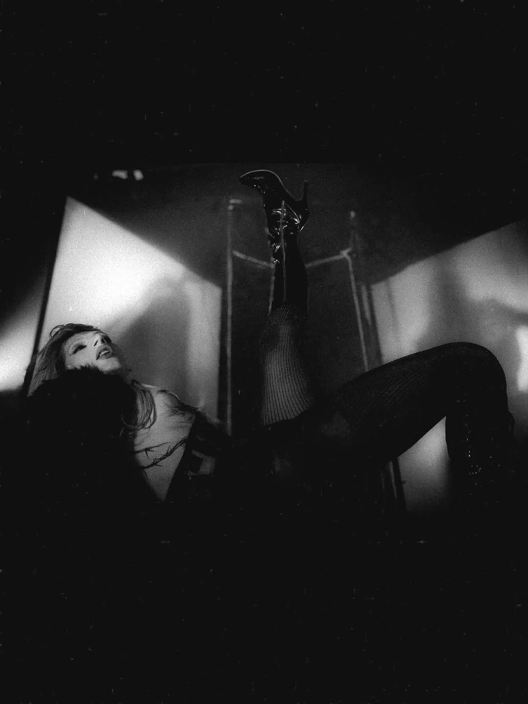 A woman with a joyful expression lying back, with one leg raised in the air and wearing high heels, black fishnet stockings, and casual clothing, in a low light setting.