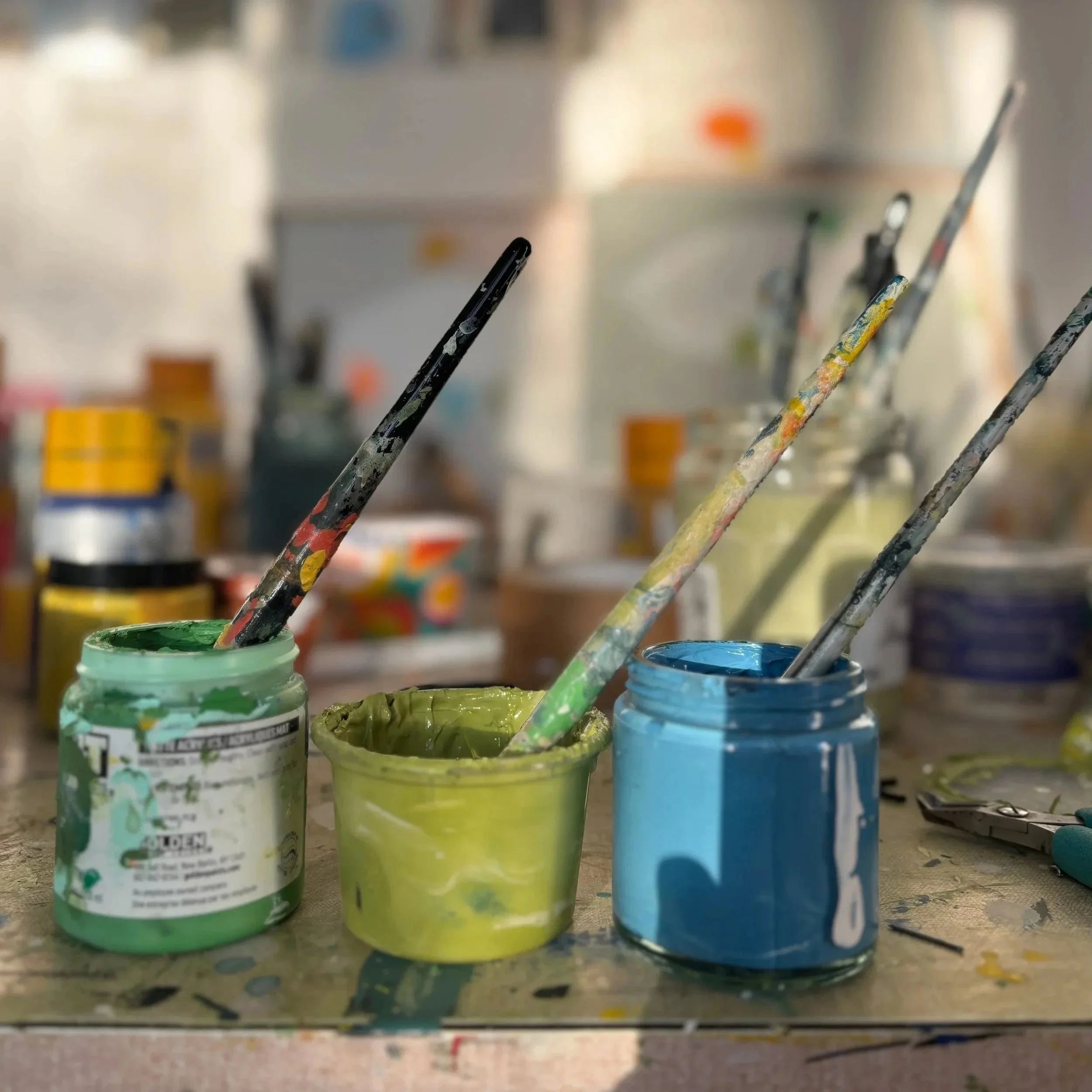 Open jars of green, yellow, and blue paint with paintbrushes inside each jar, on a paint-stained surface in an art studio or workspace.