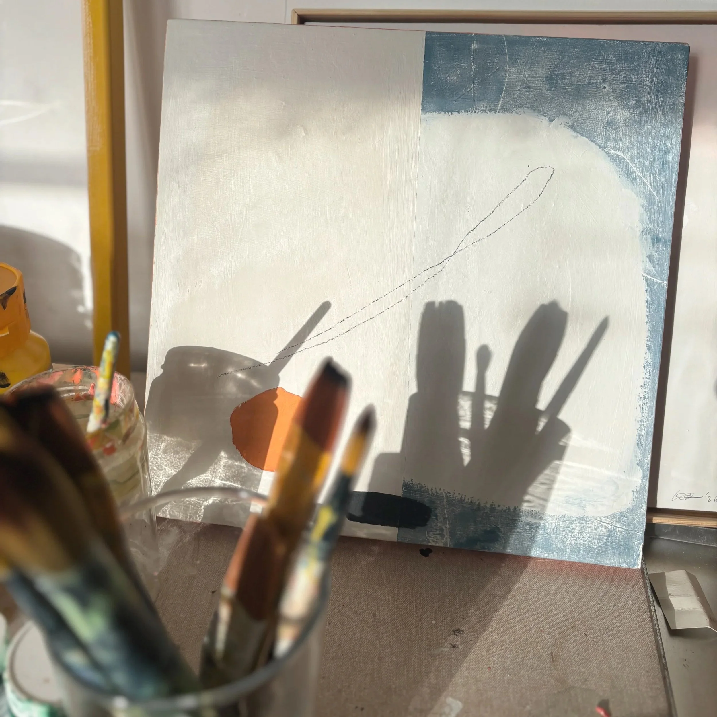Abstract painting with pastel blue, cream, and peach colors, with a faint curved line, leaning against a light-colored wall, partially obscured by the hand and paintbrush in the foreground.