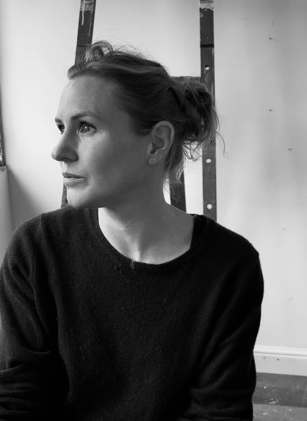 A black-and-white photo of a woman with light skin and short hair styled in a loose updo, looking to her left with a contemplative expression, wearing a dark sweater, standing in front of a plain wall.