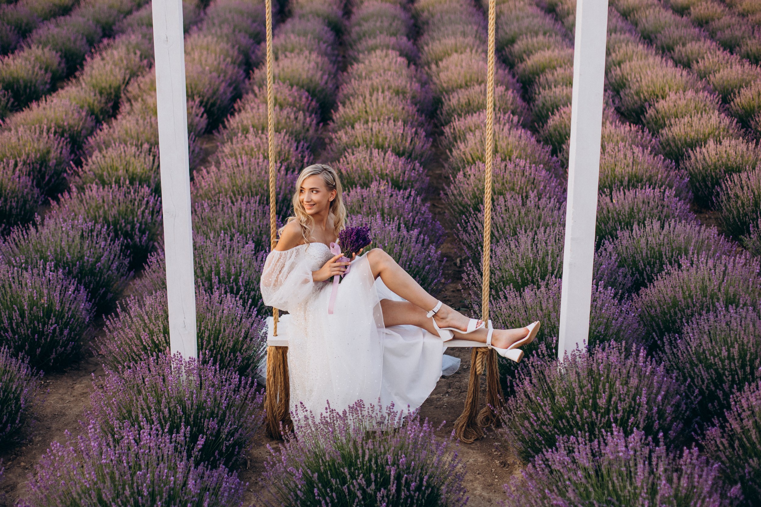 Une jeune femme en robe blanche assise sur une balançoire en bois suspendue dans un champ de lavande violet, tenant un bouquet de lavande et souriant.