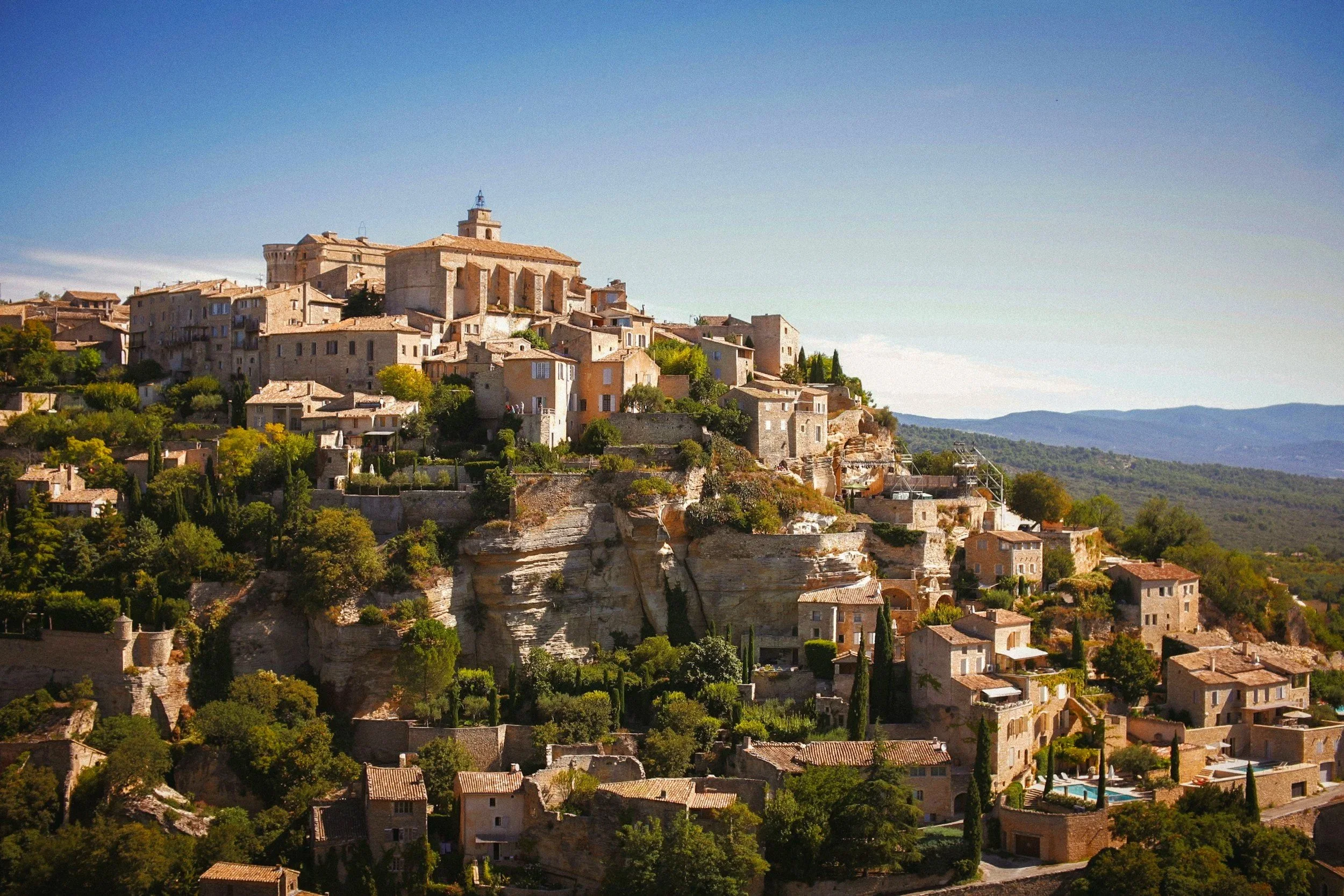 Un village perché construit sur une falaise rocheuse, avec de nombreuses maisons en pierre, entouré de verdure et de collines au loin.