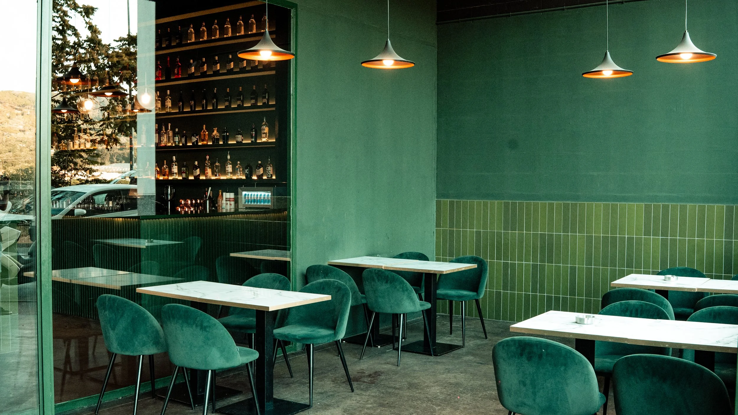 Interior de un restaurante con paredes verdes, mesas blancas y sillas verdes. Un área de bar con estantes llenos de botellas y luces colgantes.