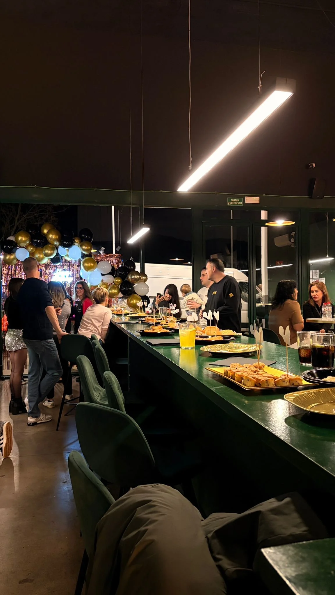 Celebración en un establecimiento con decoración de globos negros, blancos y dorados, con muchas personas conversando y disfrutando, mesas con aperitivos y bebidas, ambiente festivo.