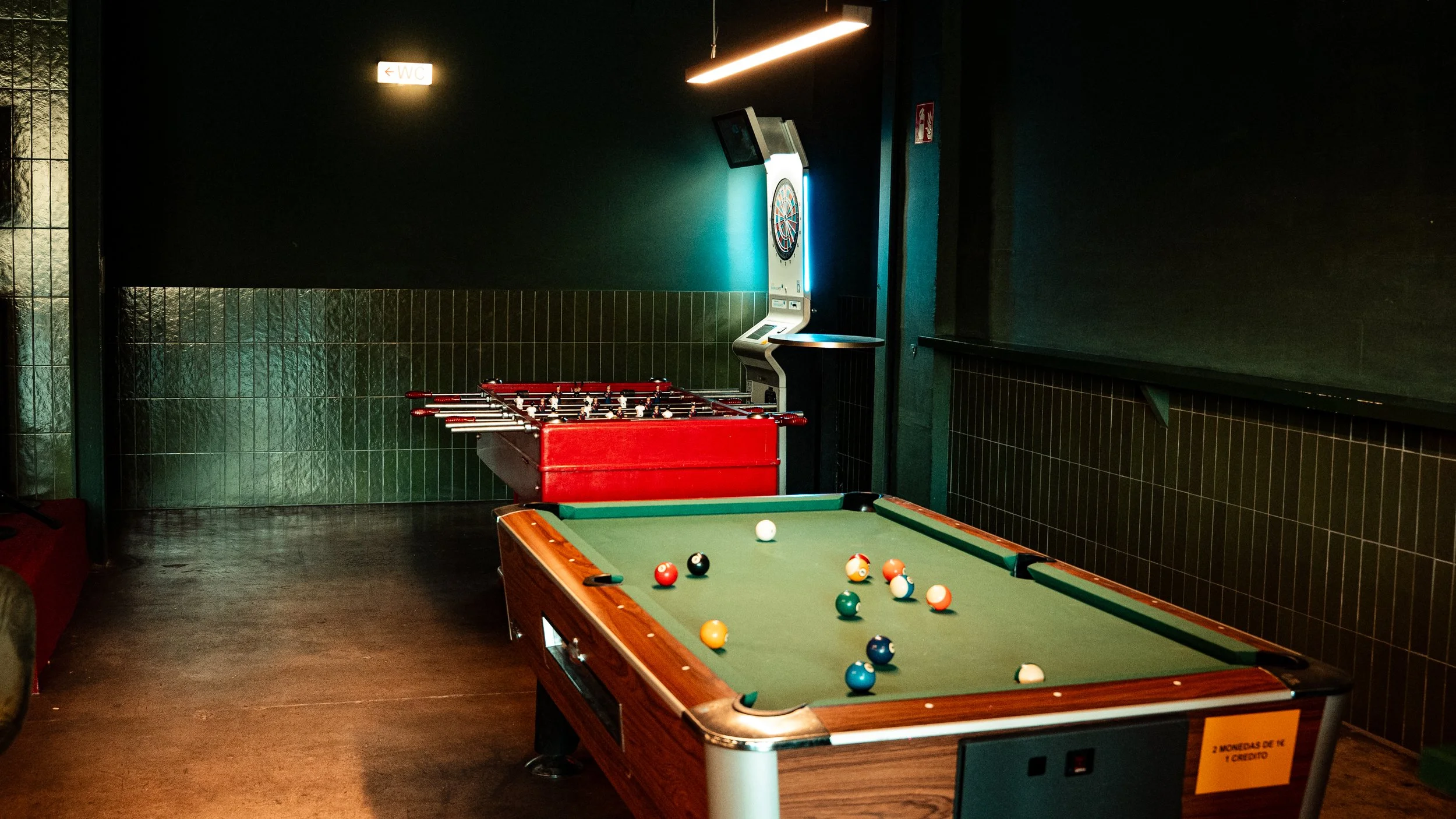 Sala de juegos con mesa de billar y máquina de dardos en fondo oscuro.