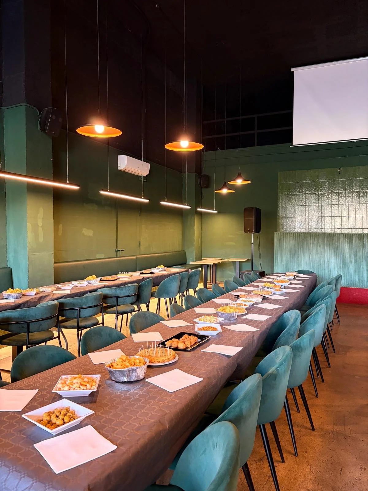 Salón de eventos con mesas largas, sillas verdes, platos de comida y decoración moderna, preparado para una fiesta o reunión.