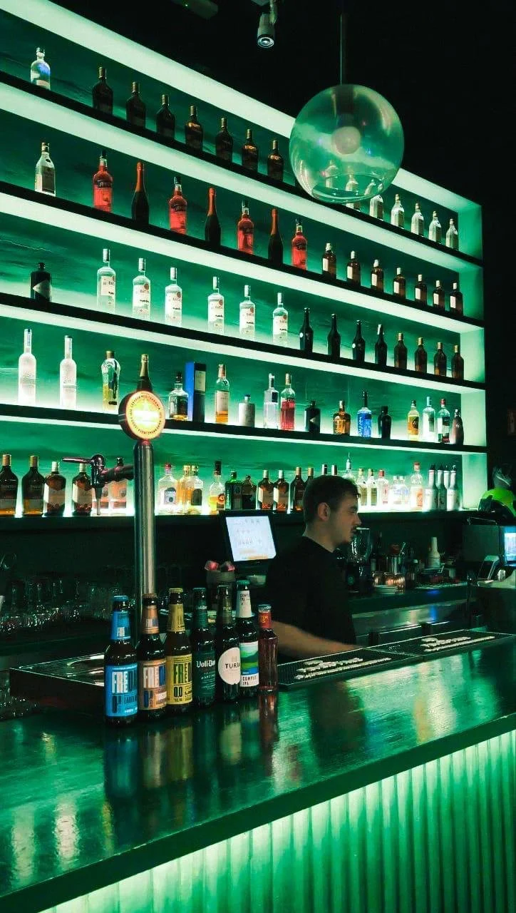 Bar con estanterías de botellas de bebidas alcohólicas, luces de fondo verdes, bartender atendiendo, ambiente nocturno