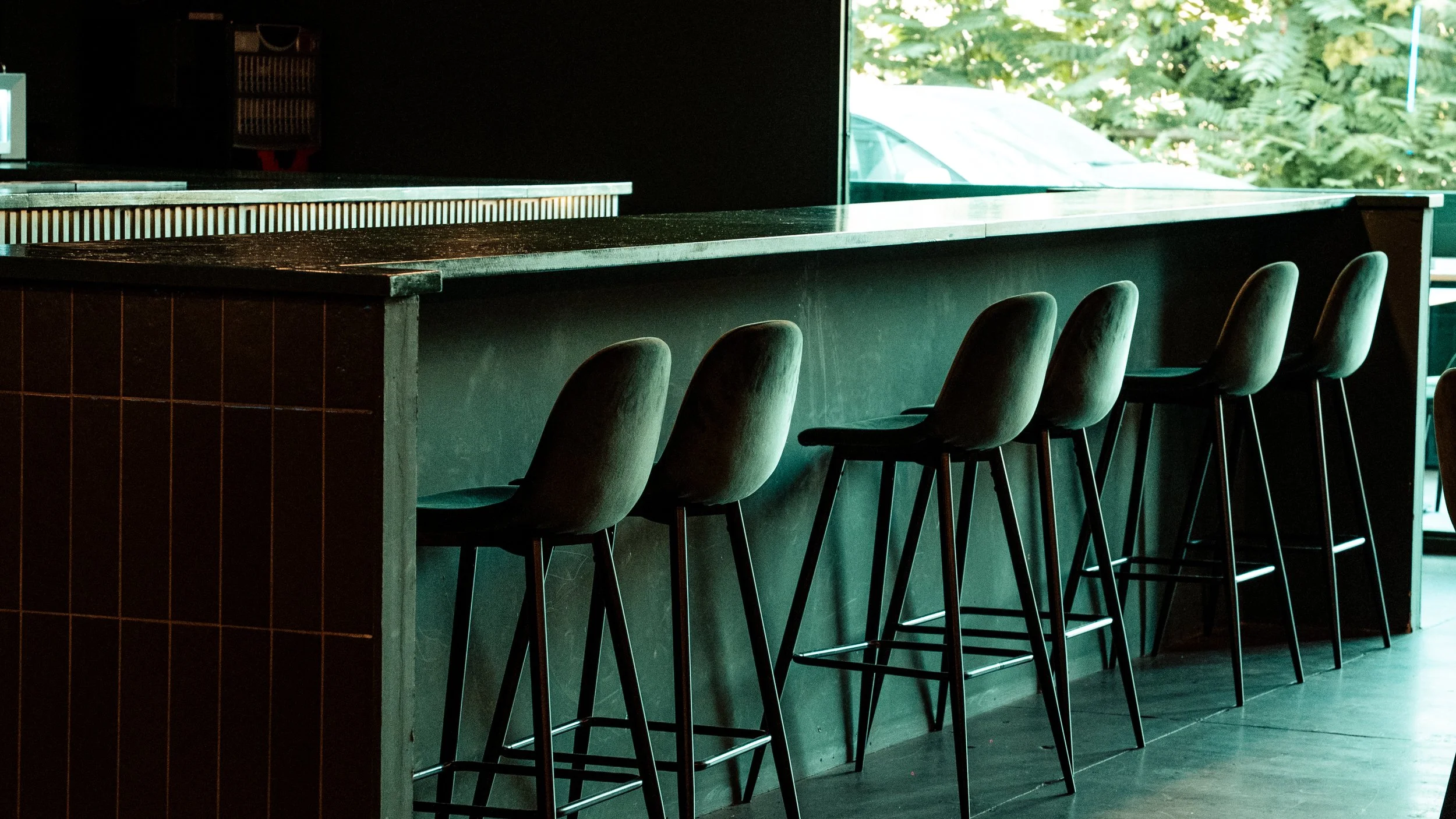 Bar con seis sillas altas de respaldo acolchado y patas metálicas, y una superficie de mostrador de madera oscura, en un establecimiento con amplias ventanas y vista a árboles verdes