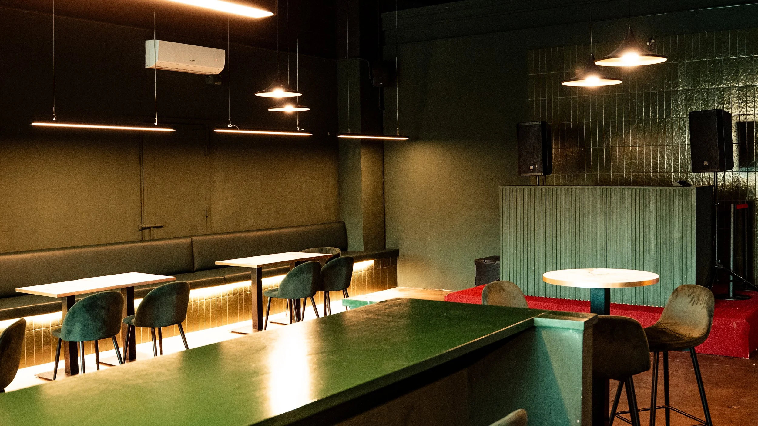 Interior de un bar o restaurante con mesas, sillas y luces modernas, paredes oscuras y un ambiente acogedor, espacio para música en vivo con altavoces y un pequeño escenario.