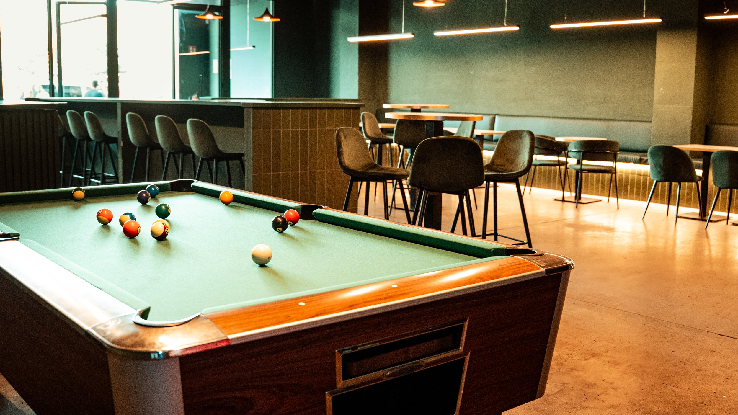 Una mesa de billar en un café o bar con sillas y mesas en el fondo, iluminación cálida y grandes ventanas con luz natural.