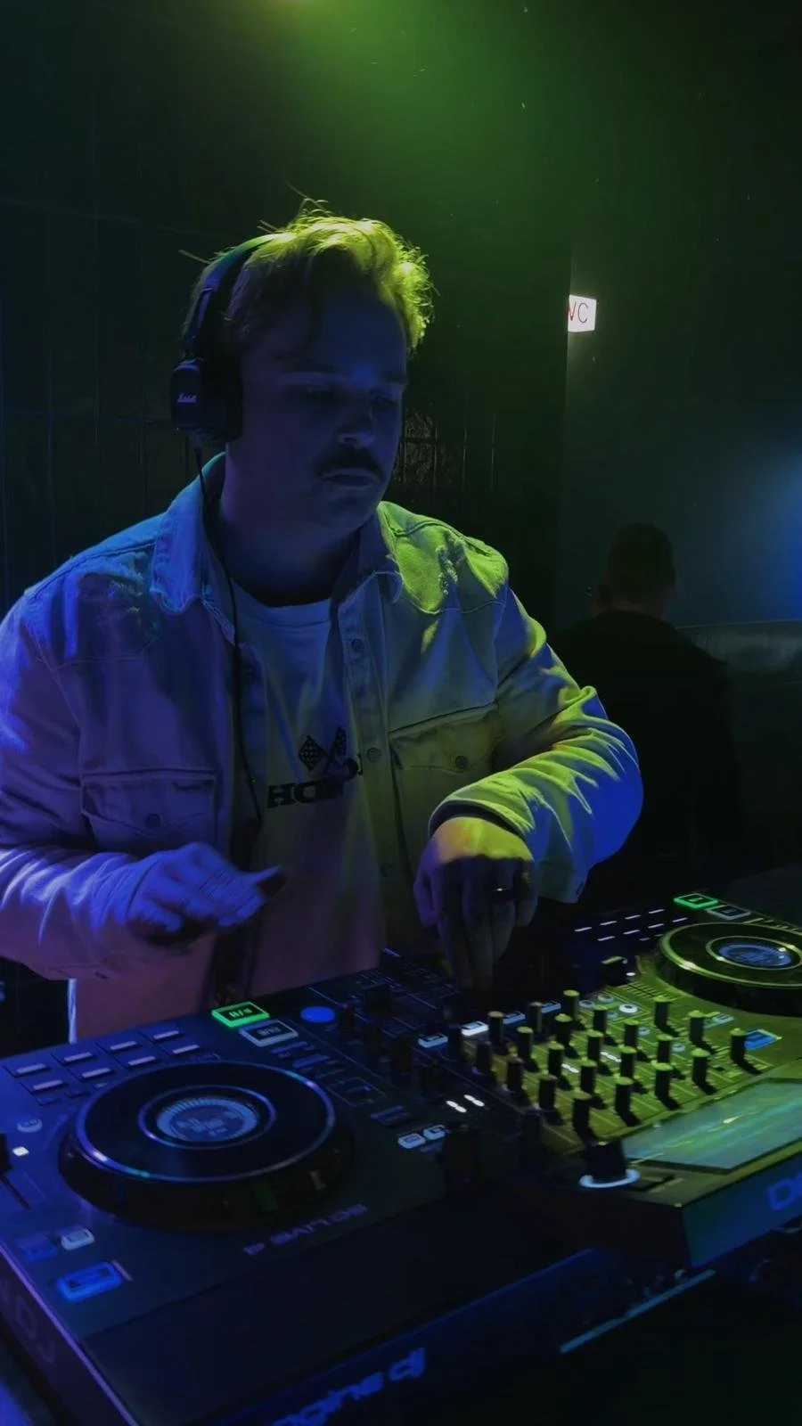 Joven DJ con auriculares mezclando música en un club con iluminación de neón verde y azul.