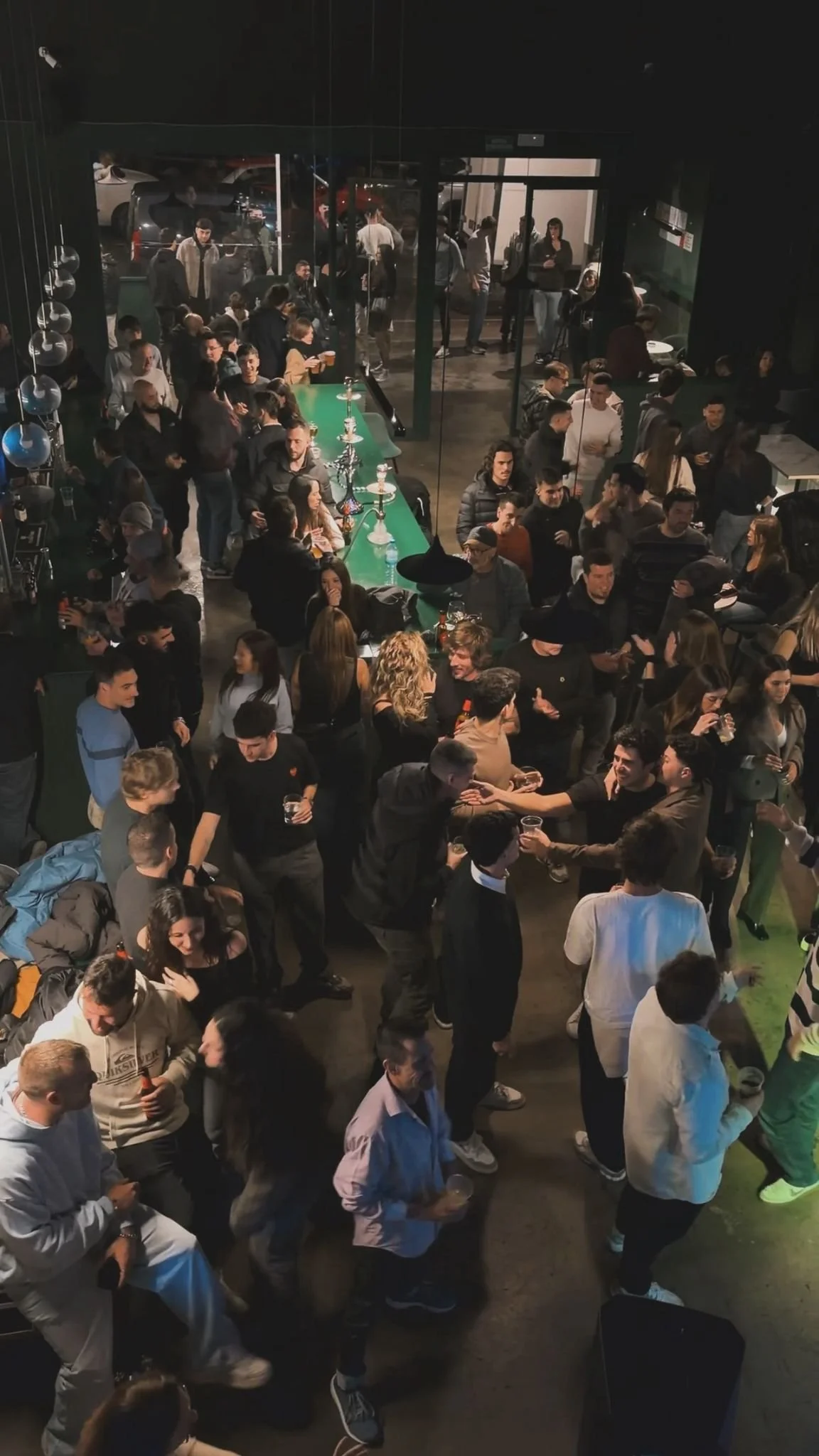 Vista de una fiesta o reunión social dentro de un lugar cerrado, con muchas personas conversando y disfrutando. Hay una barra verde en el centro con varias tortas, y algunas personas están en la entrada del lugar, que tiene puertas de vidrio y autos estacionados afuera.