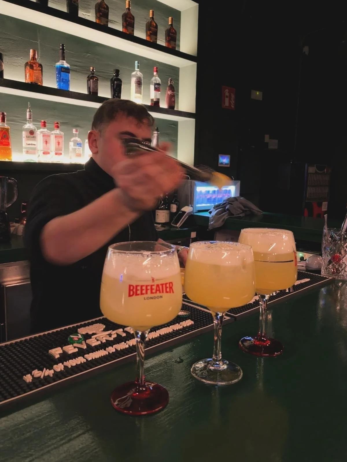 Barman preparando cócteles en un bar, delante de tres copas con bebidas amarillas y espuma en la parte superior.