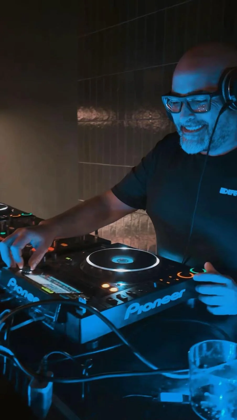 Un DJ con gafas, barba y camiseta negra, sonriendo mientras mezcla música en un equipo Pioneer en un ambiente oscuro con iluminación azul.