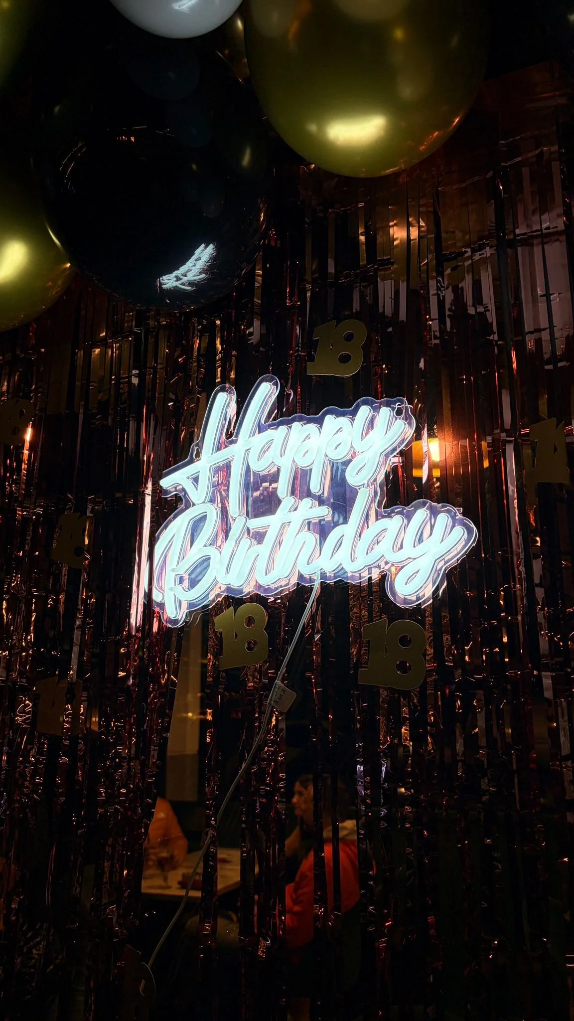 Decoración de cumpleaños con globos dorados y negros, y letrero de neón que dice 'Happy Birthday' en inglés, en un fondo de cintas y adornos dorados con el número 18.