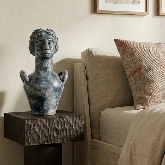 Statue en céramique stylisée d'une tête humaine avec des bras, placée sur une table de nuit en bois à côté d'un lit.