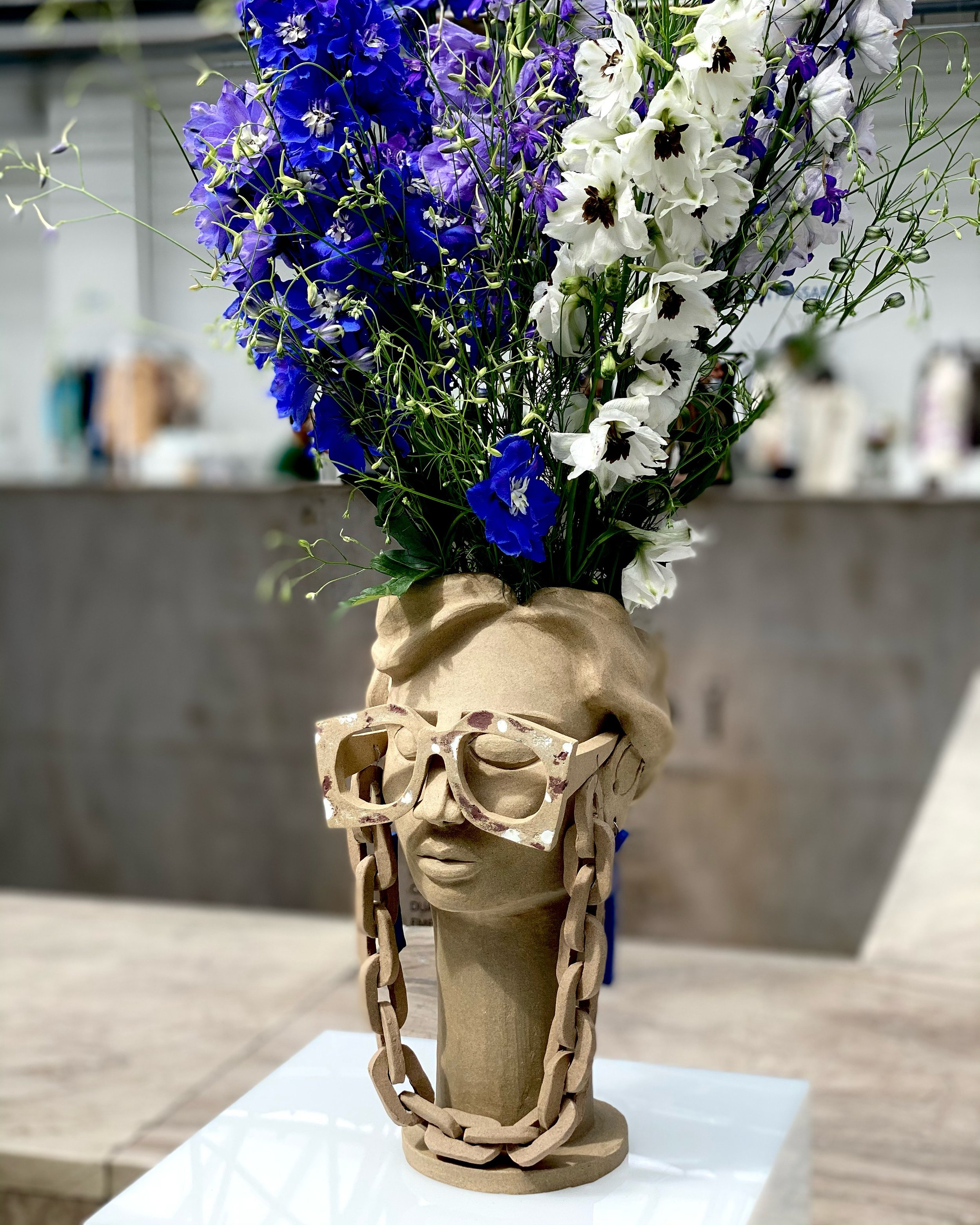 A wooden sculpture shaped like a human face wearing glasses, used as a stand for a bouquet of purple and white flowers. The sculpture is placed on a white surface in an indoor setting.