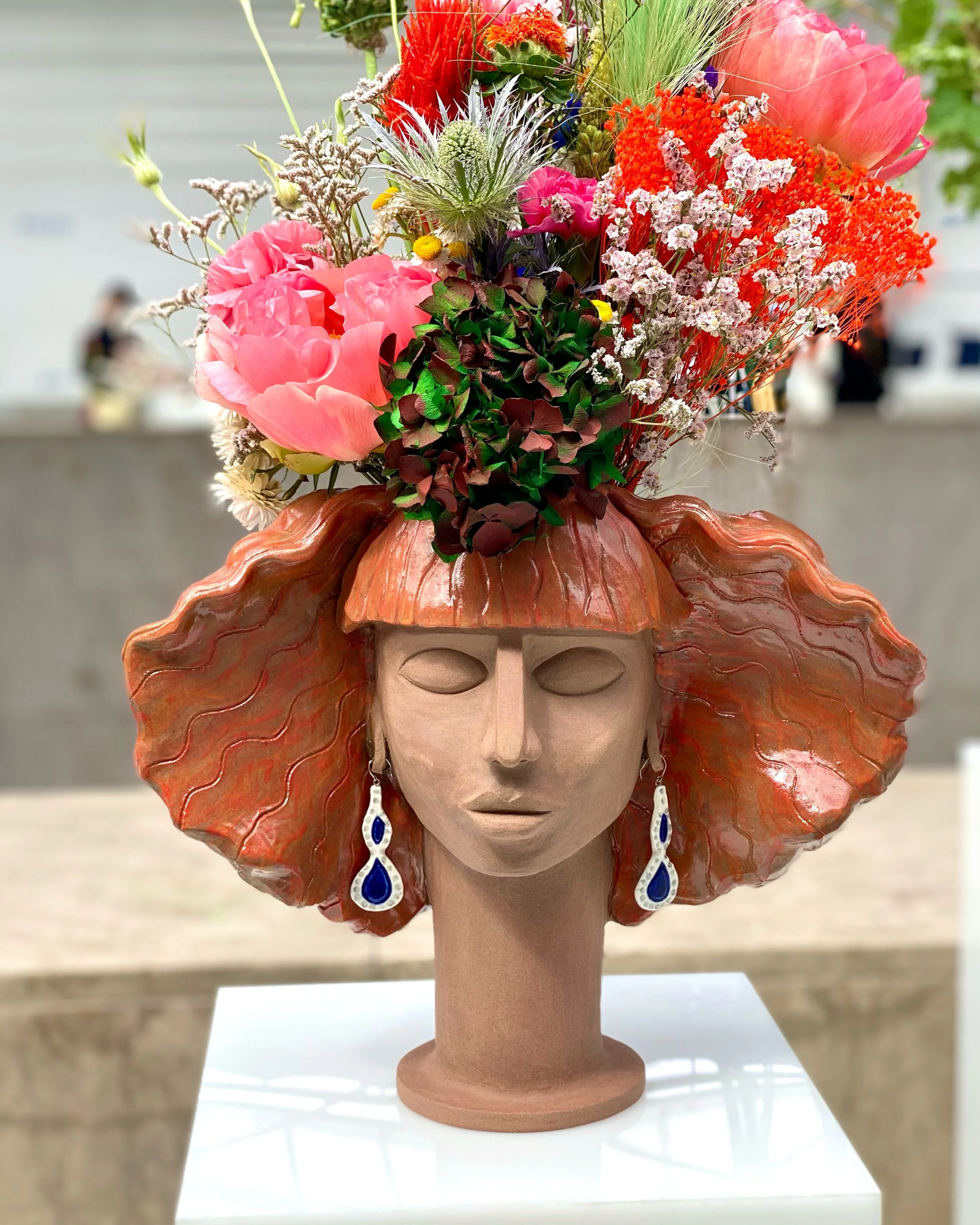 A sculpture depicting a woman's head wearing a large orange seashell-shaped hat, adorned with multicolored flowers, and sporting blue and white earrings.
