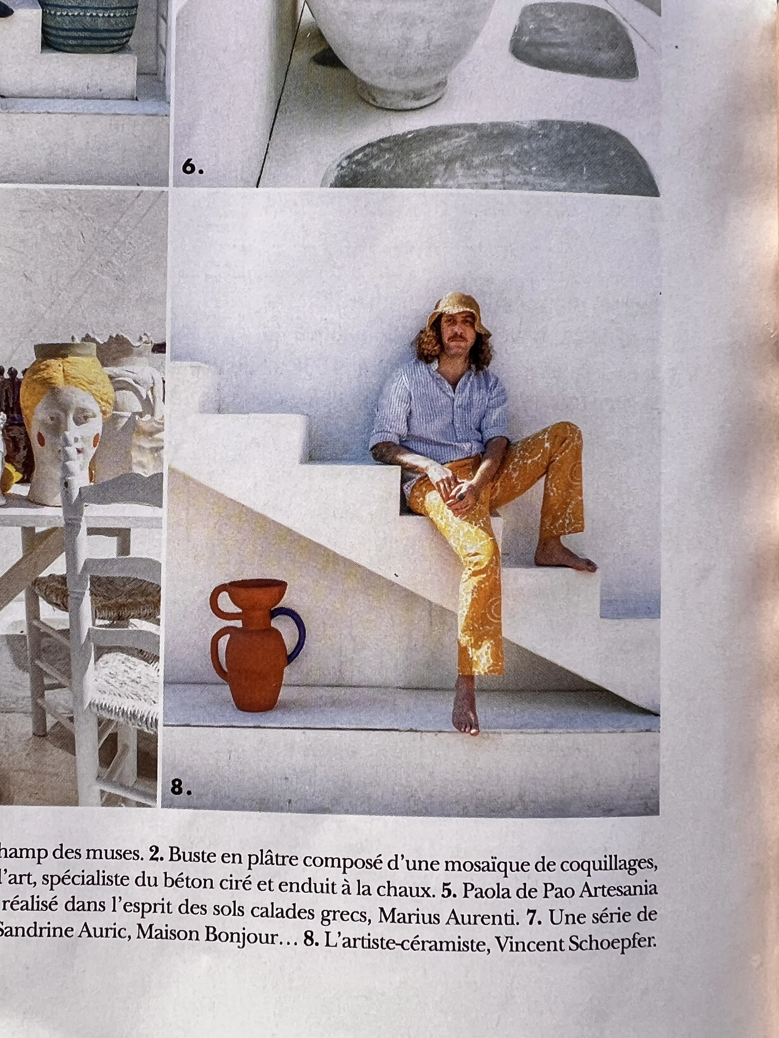 A person with curly hair wearing a hat, sitting on white stairs, wearing patterned orange pants and a striped shirt, in a minimalist setting with white ceramic objects and terracotta vases.