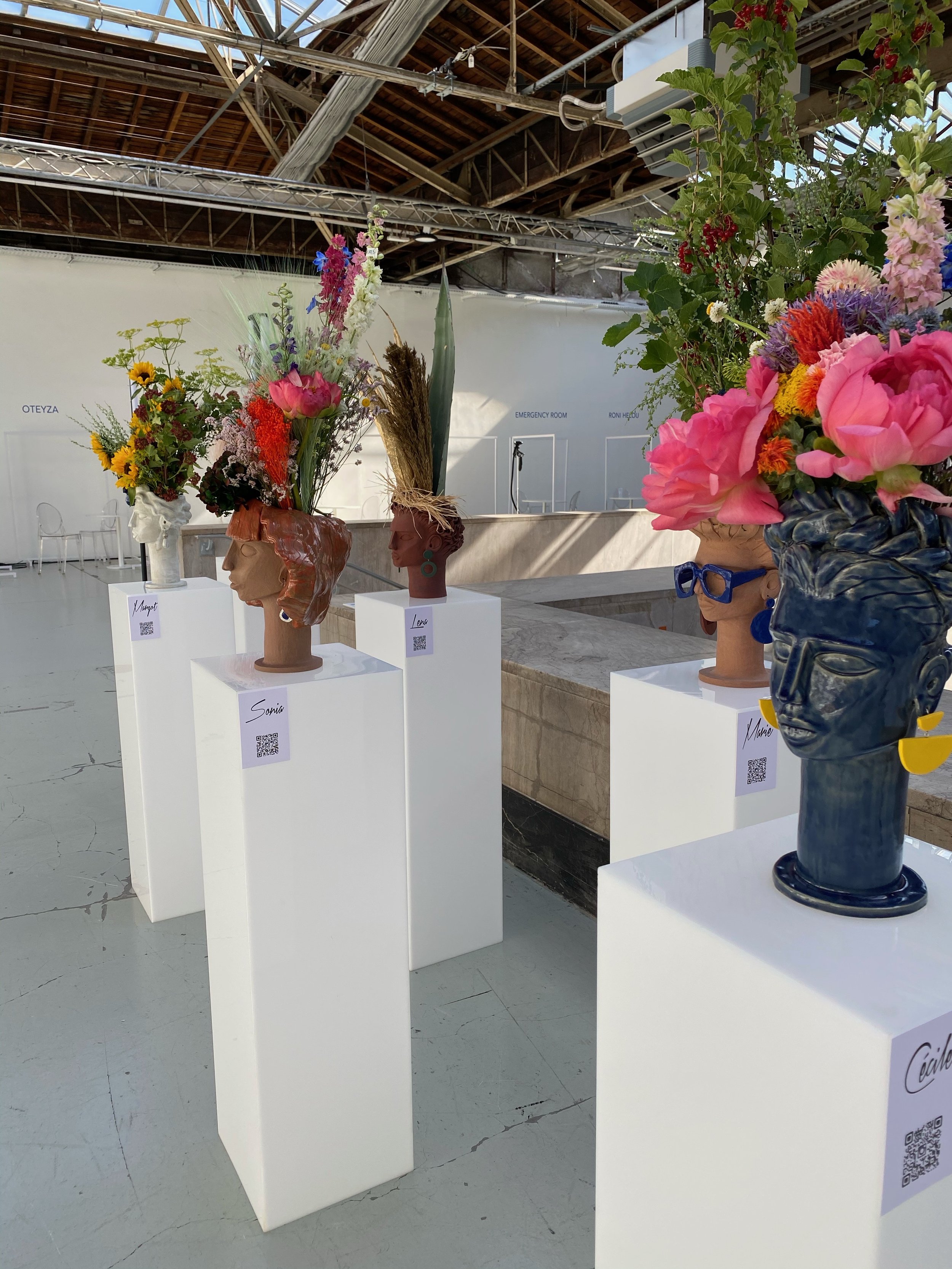 An exhibition of ceramic sculptures of women's heads, adorned with colorful floral arrangements, in an indoor exhibition space.