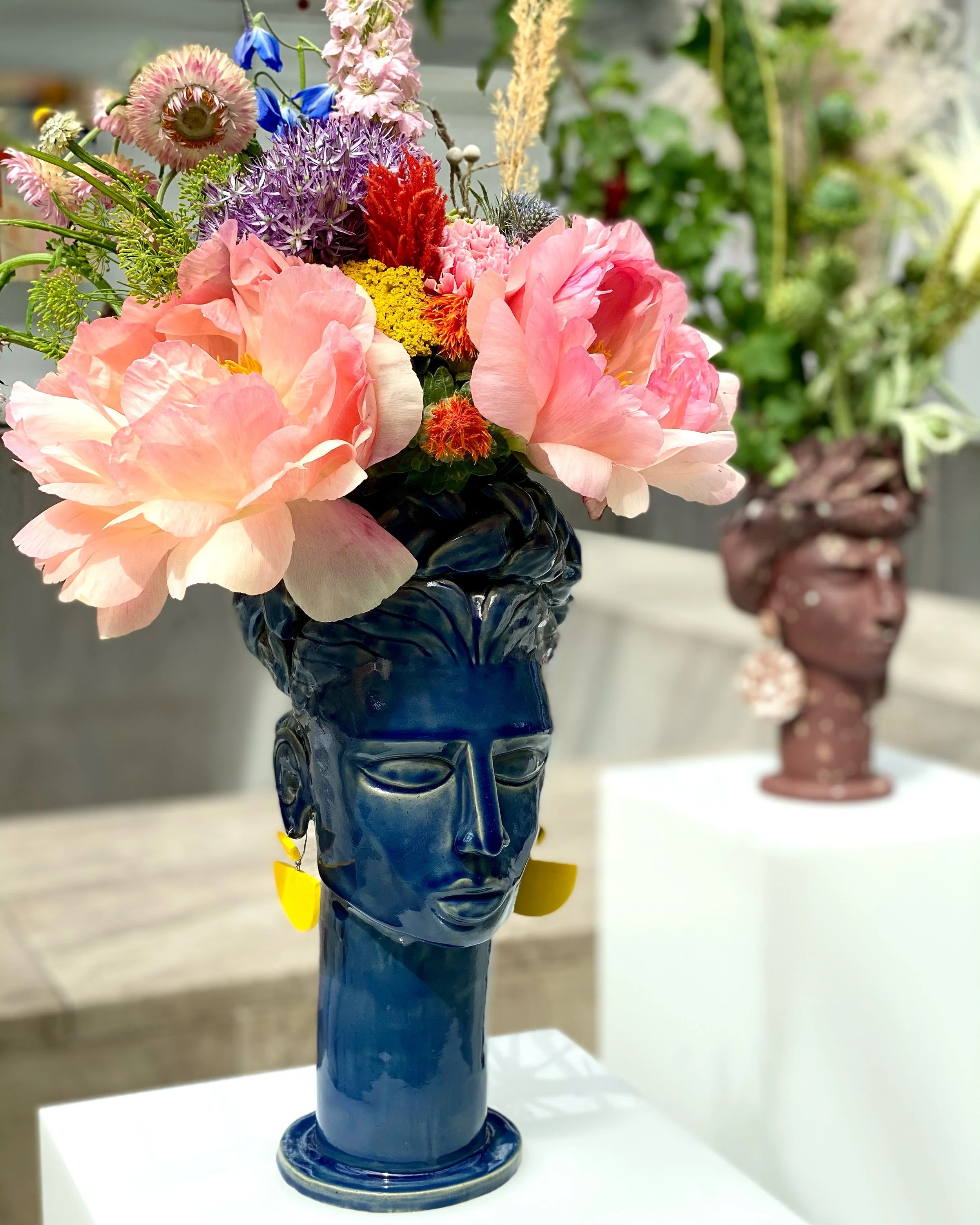 A blue ceramic vase shaped like a face, featuring colorful flowers—including large pink peonies, purple flowers, pink flowers, and a few yellow flowers—arranged in an inner setting.