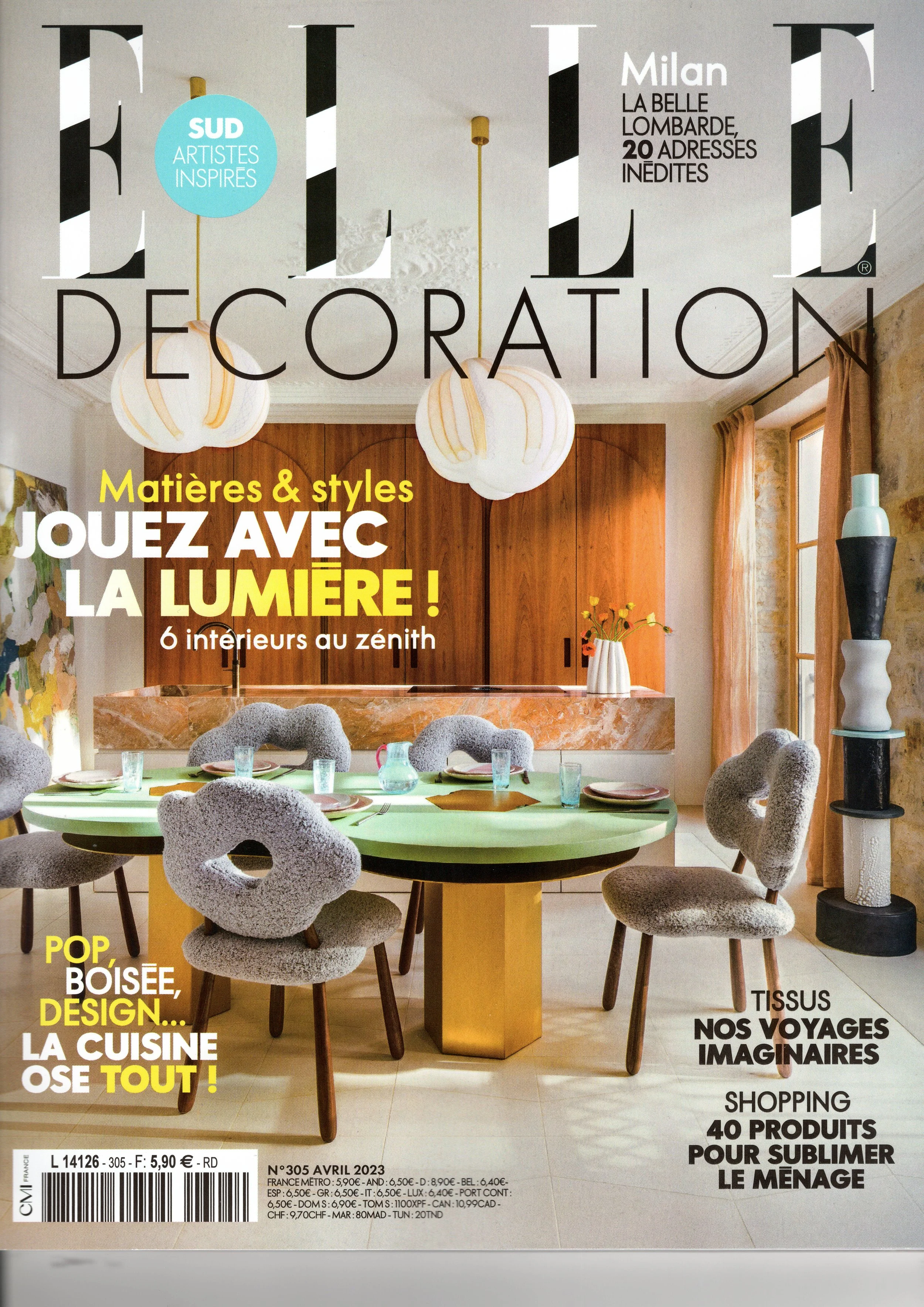 A modern and elegant room featuring a green round table, gray fabric chairs, a wooden cabinet in the background, pendant lights hanging from the ceiling, and a variety of decorative items. Text in French on the cover of Elle Décoration magazine.