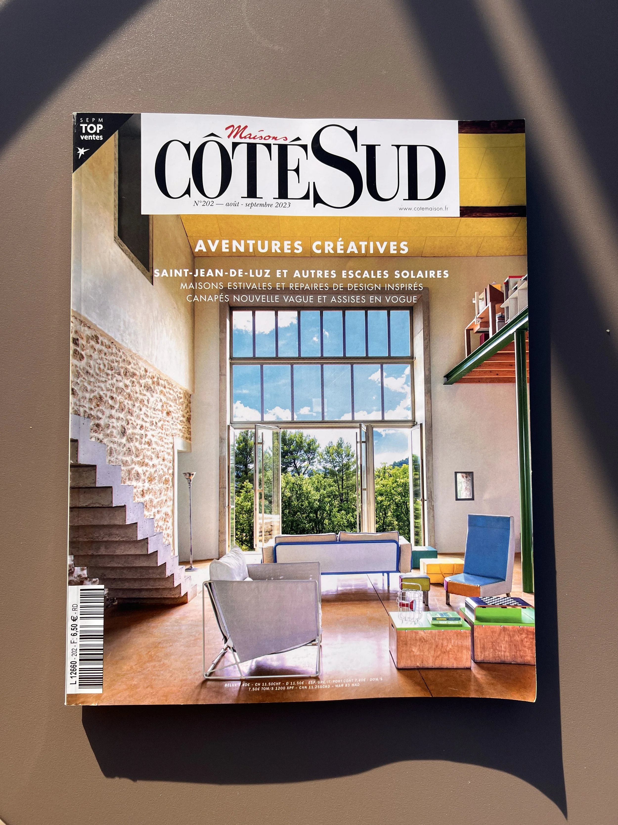 Magazine cover featuring a bright living room with a large window, colorful armchairs, wooden tables, a stone staircase, modern decor, and a view of the natural landscape outside.