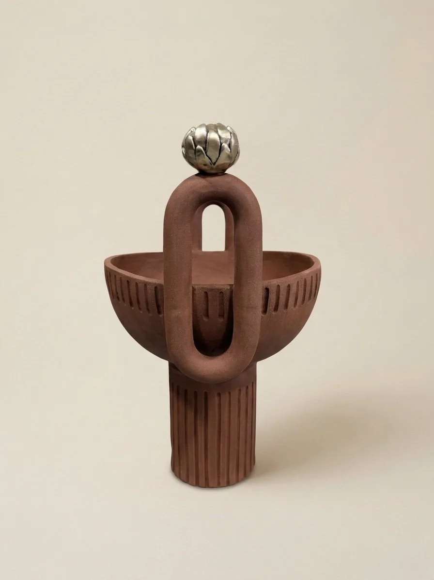 Une sculpture en céramique représentant un objet abstrait avec une base cylindrique, un bras en forme de boucle, une coupe ou récipient, et une décoration en forme de tête ou de fruit en haut, en métal argenté.