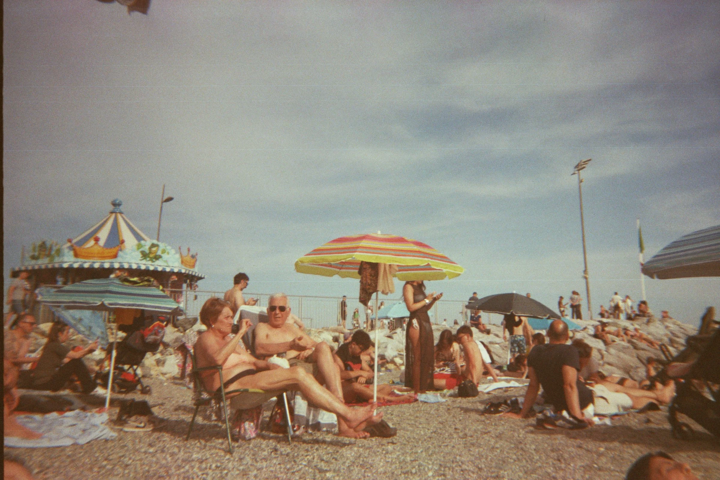 People relaxing on a crowded beach with umbrellas and a carousel in the background.