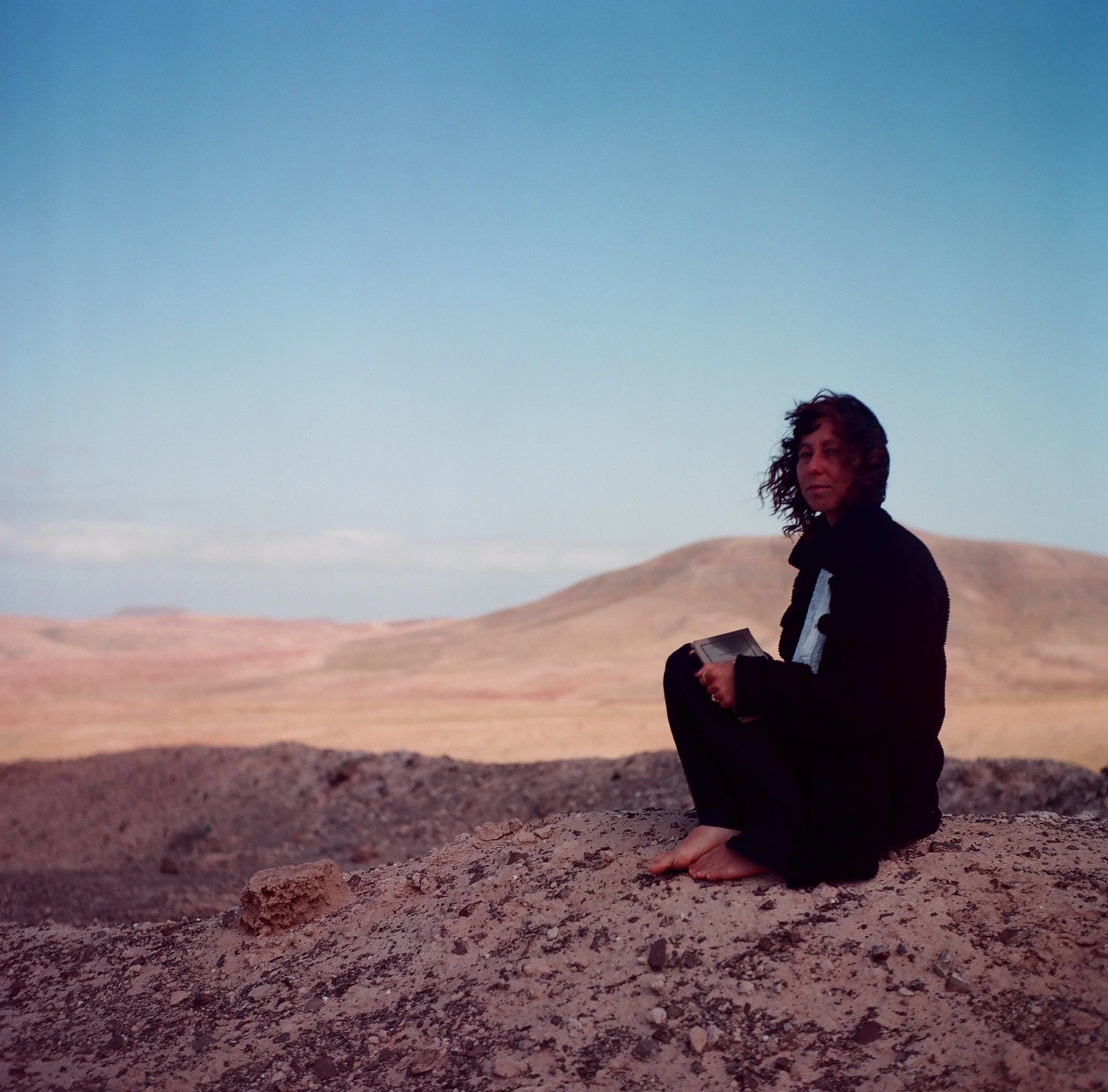 A woman with curly hair sitting barefoot on a rocky desert landscape, holding a tablet, with hills and a blue sky in the background.
