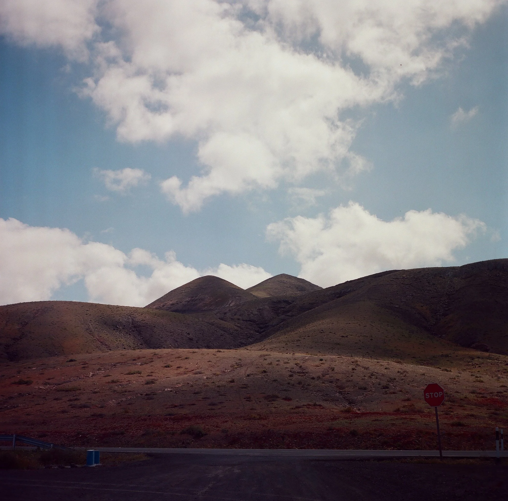 Dry, hilly landscape with sparse vegetation, a stop sign, and a road at the foreground under a partly cloudy sky.