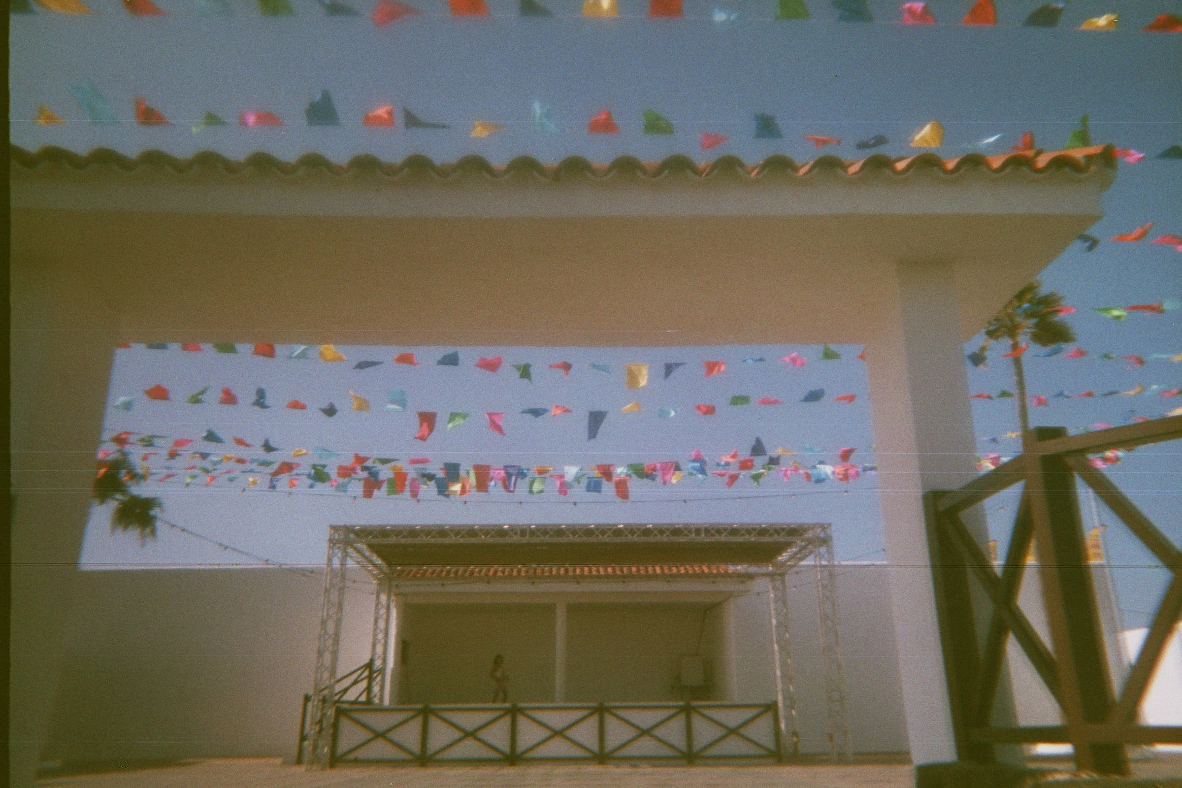 Colorful triangular flags hanging across an outdoor space with a clear blue sky, a stage with a metal frame, and a person standing on a balcony.