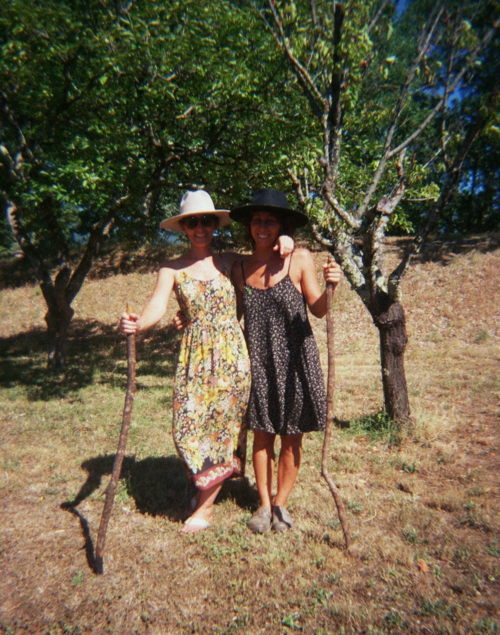 Two women standing outdoors next to a tree, wearing wide-brimmed hats and dresses, smiling, with a sunny and dry environment in the background.