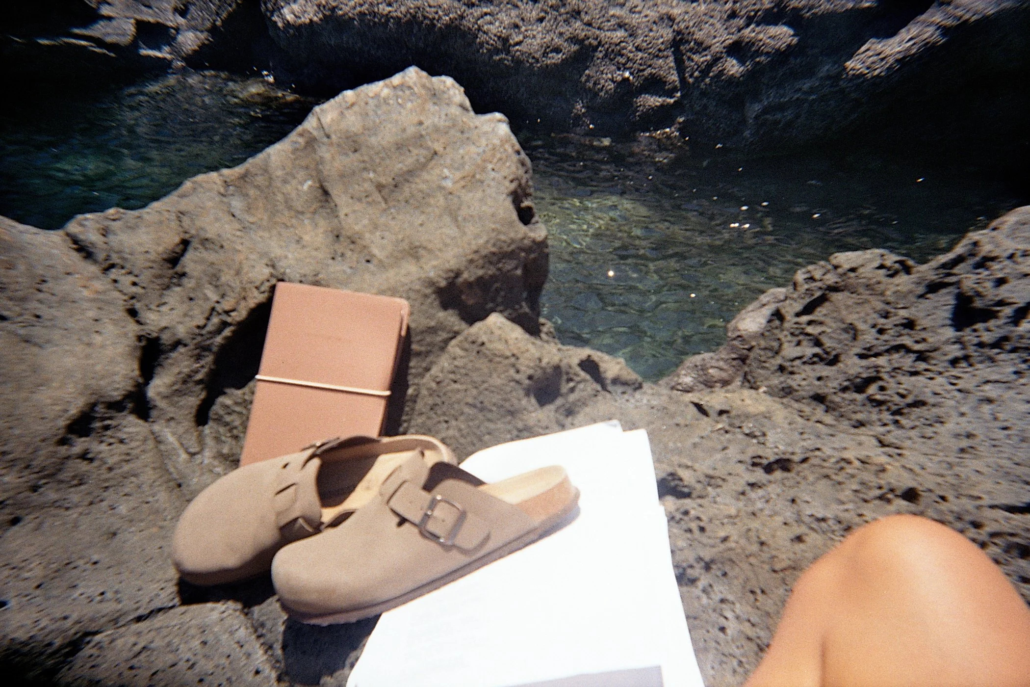 Beach scene with rocks, a pair of beige sandals, a notebook, and a white piece of paper on rocks near water
