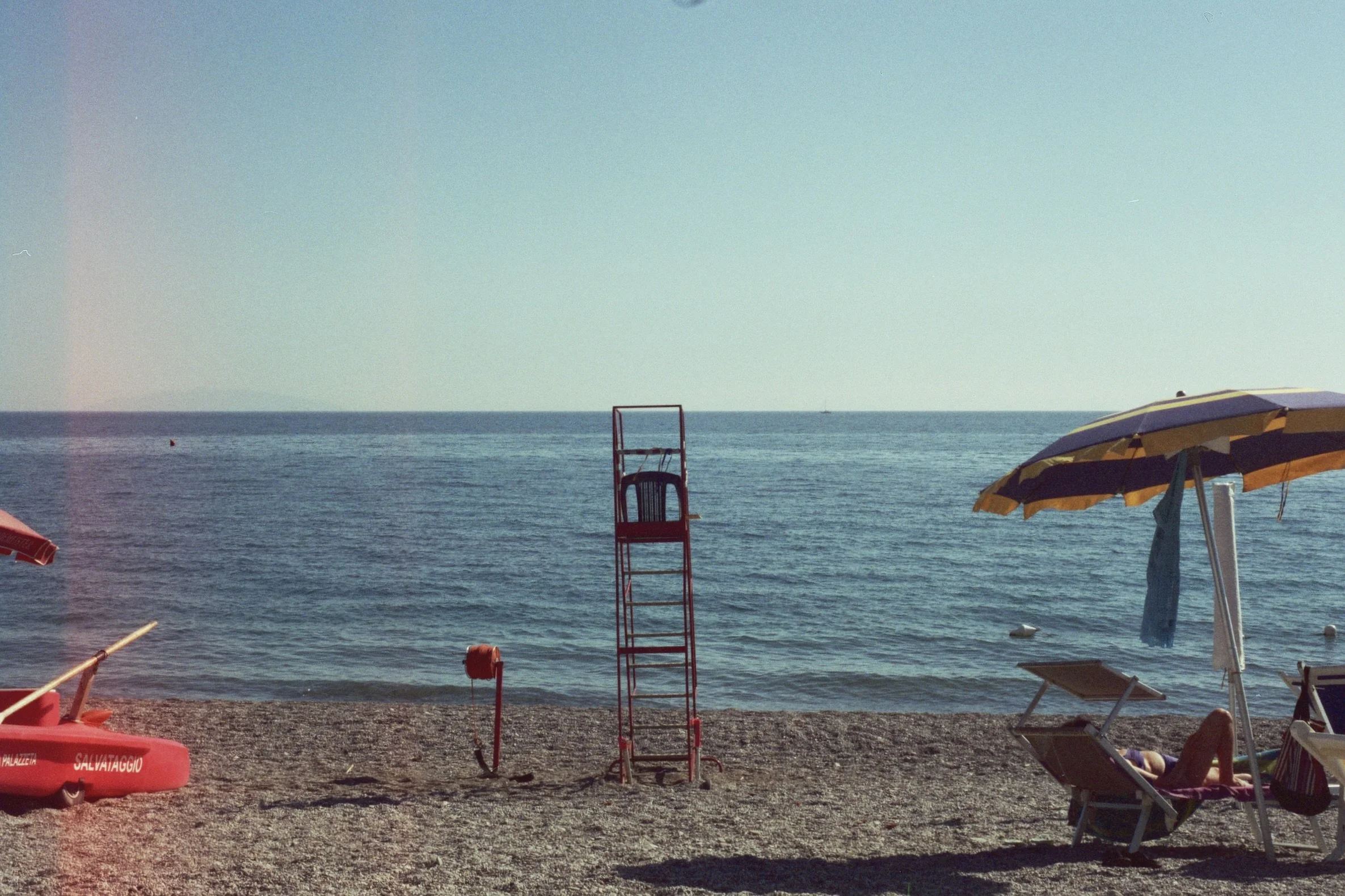A beach scene with a lifeguard chair, umbrellas, and lounge chairs on a pebbly shore, with the ocean and a clear sky in the background.
