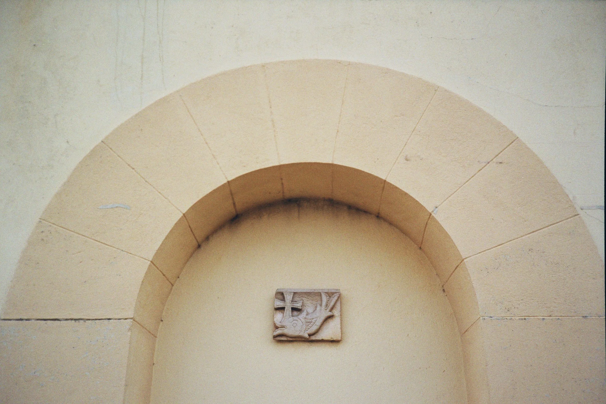 A small stone plaque with a carved Christian cross and an eagle facing to the right, mounted inside a beige archway on a light wall.