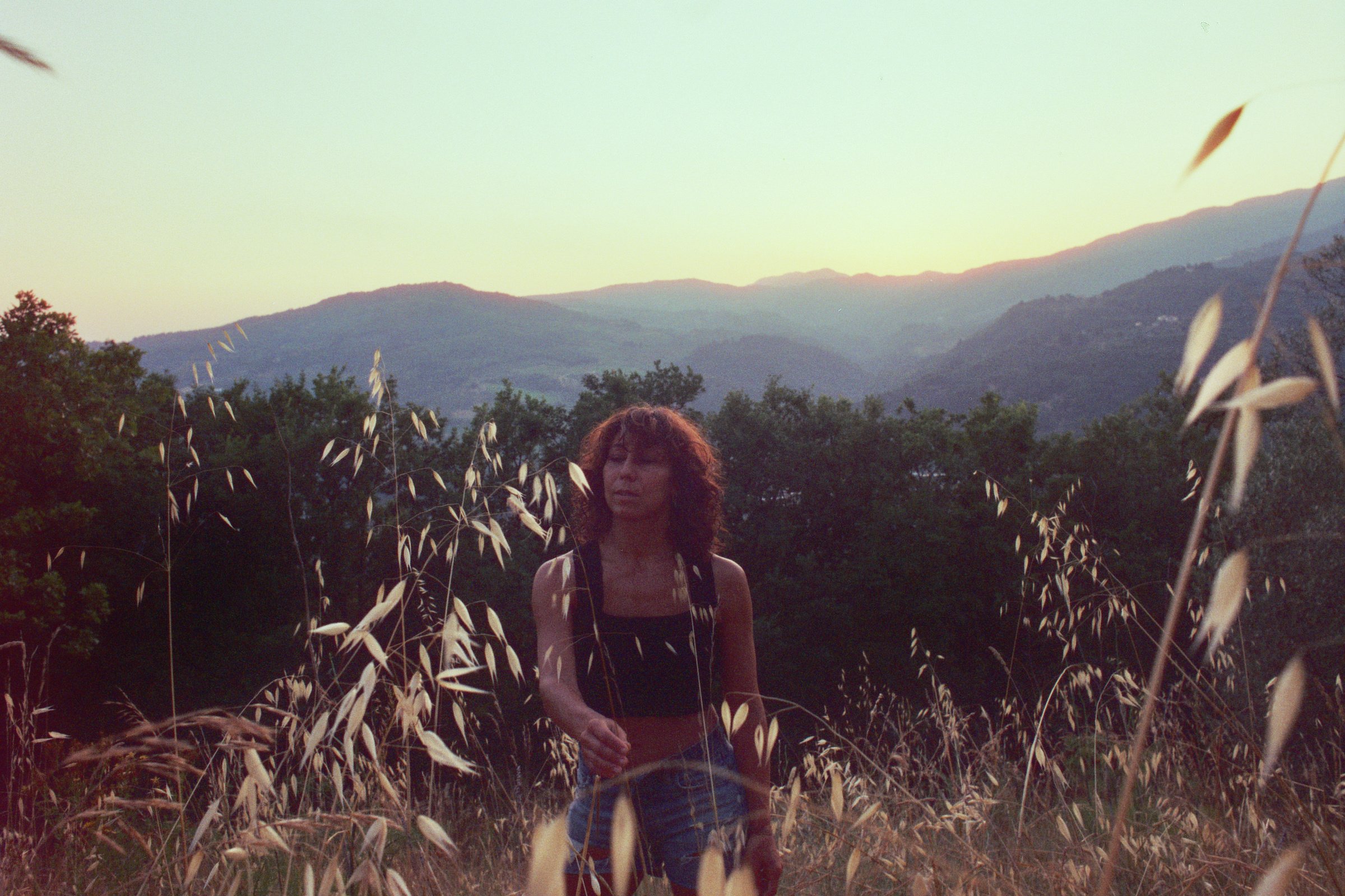 A young woman with curly hair standing in a grassy field during sunset, with mountains and green trees in the background.