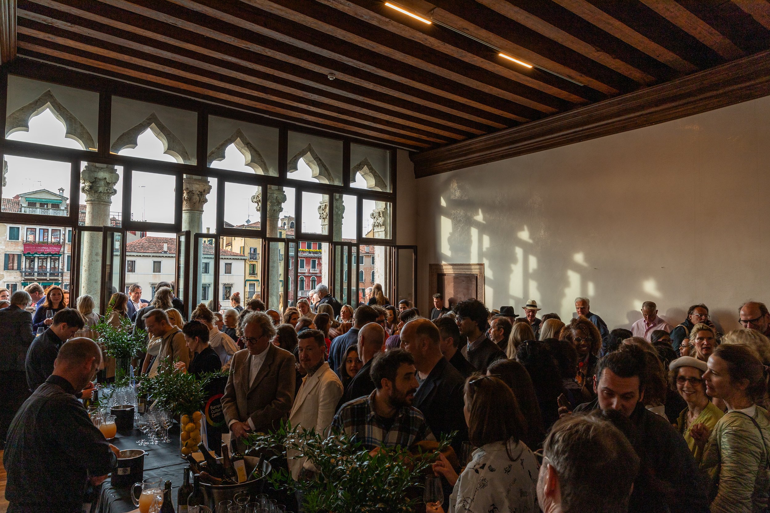 Crowd of people gathered inside a historic building with large arched windows and columns, enjoying a social event or reception.