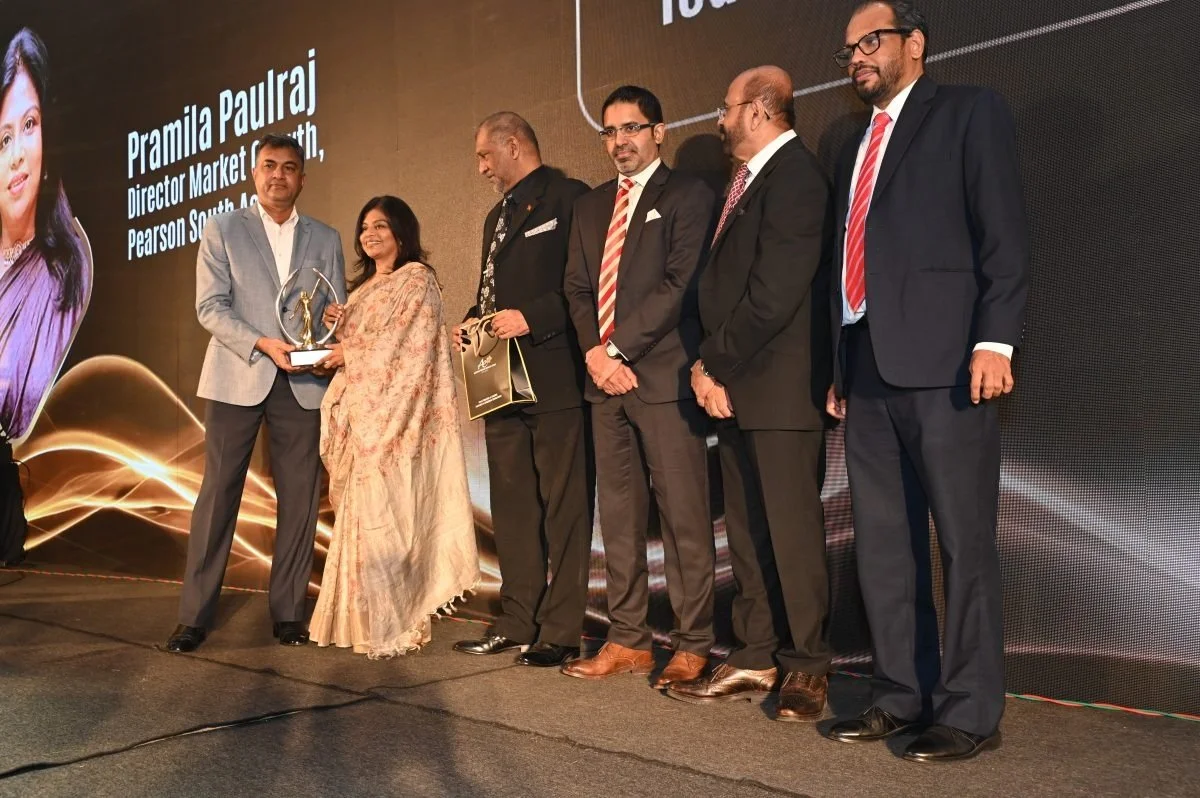 Group of six people on stage during an award ceremony, with large screen displaying a woman's face and the name Pramila Paulraj, in formal suits and traditional attire.