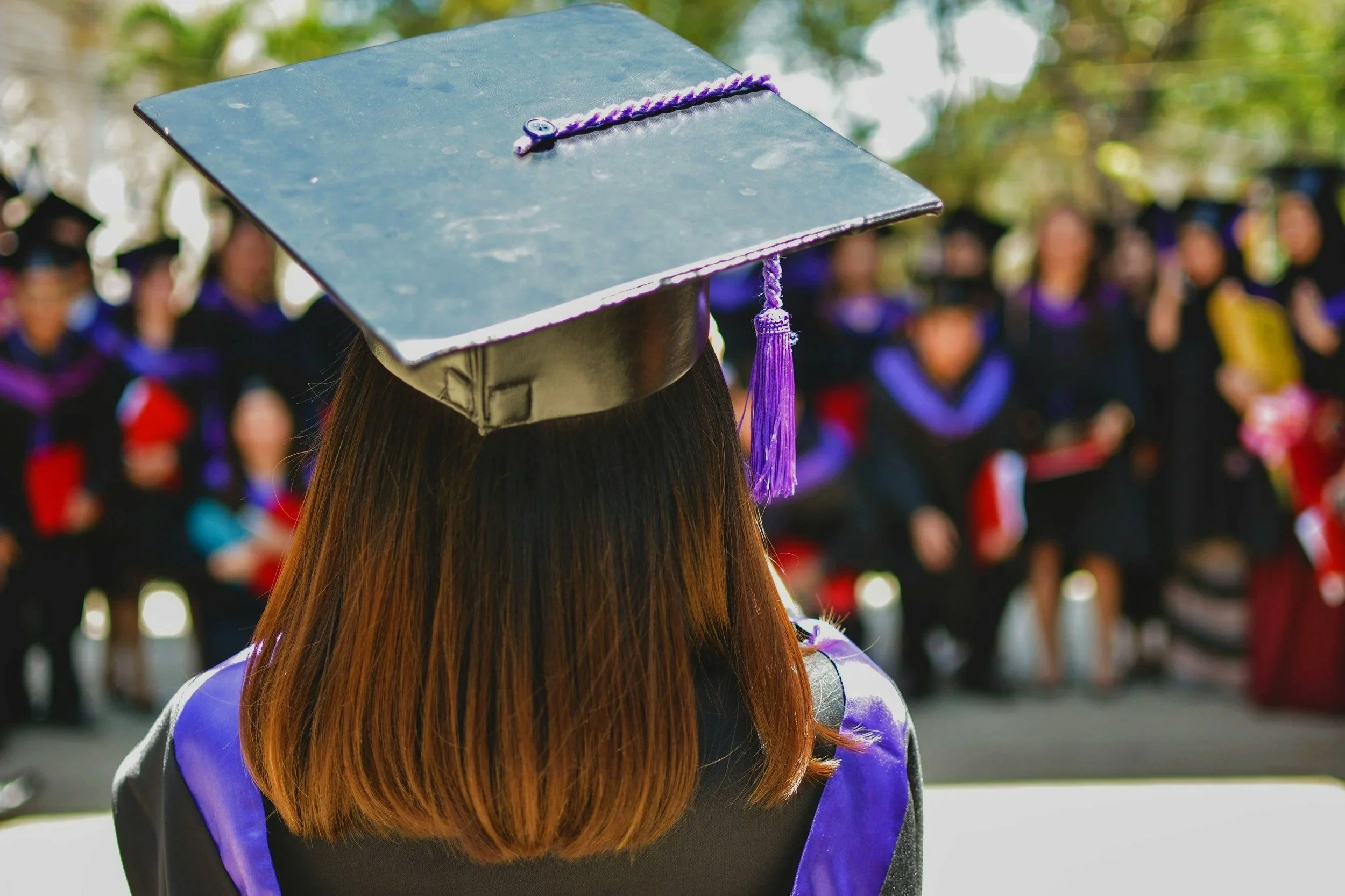 A graduate wearing a black cap and gown with purple accents, seen from behind during a graduation ceremony, with other graduates in the background.