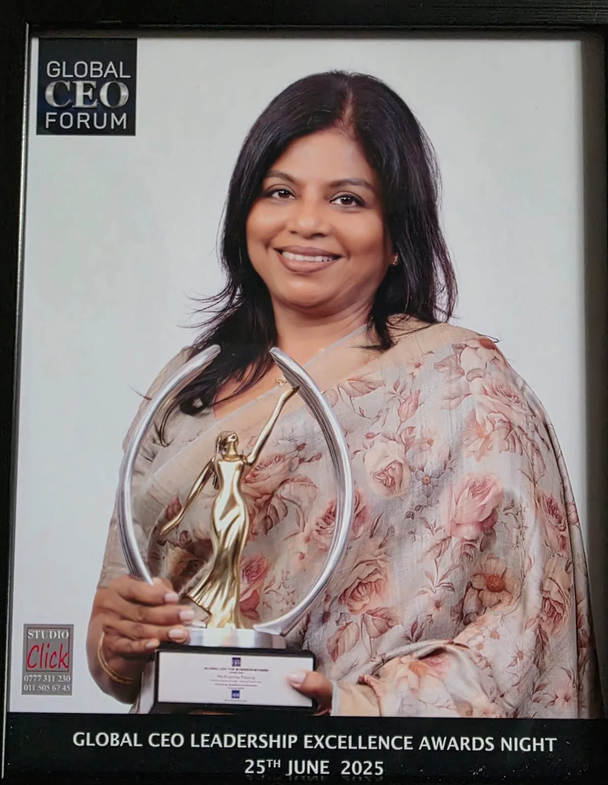 A woman with shoulder-length black hair, wearing a light-colored floral dress, holding a trophy with a gold figure of a woman and the text "Global CEO Leadership Excellence Awards Night" in the background, dated June 25, 2025.