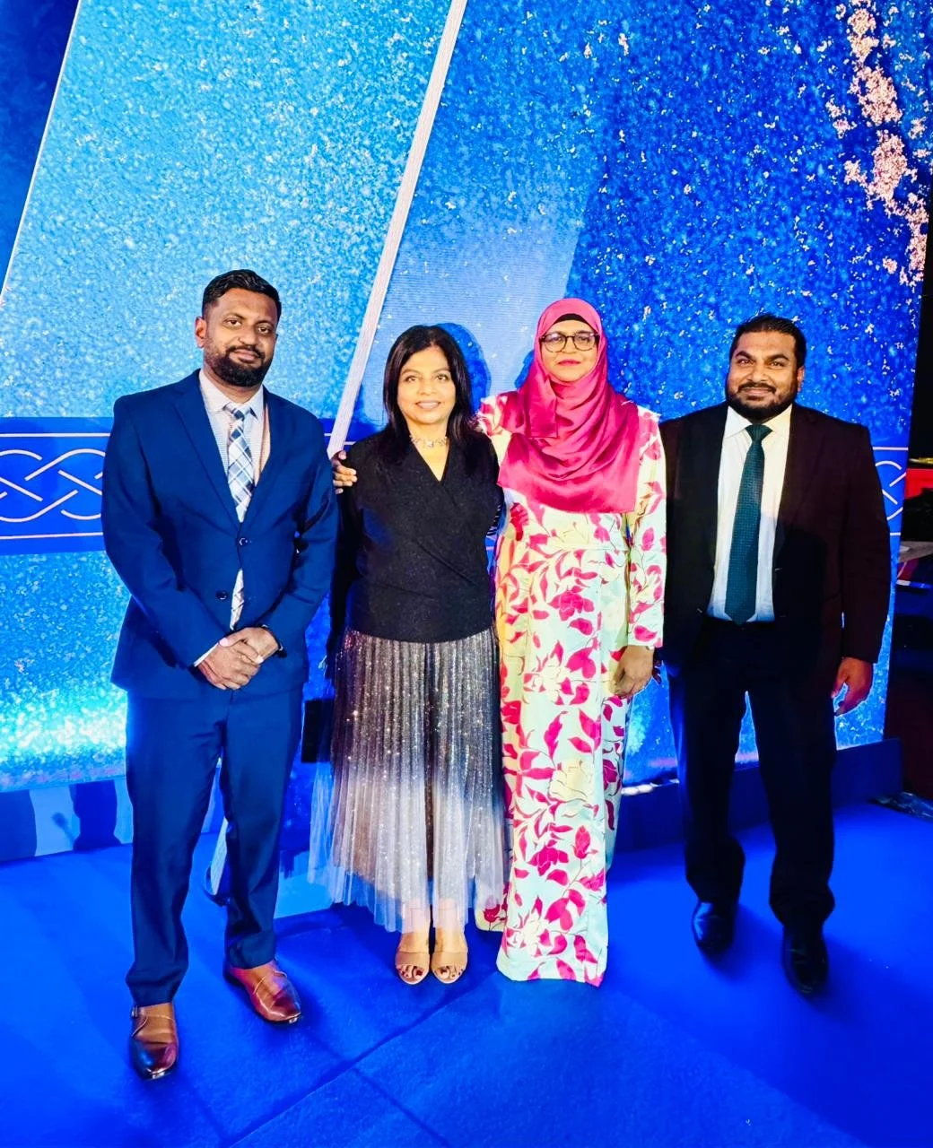 Four people standing together dressed in formal and traditional attire against a blue glittery photo wall.