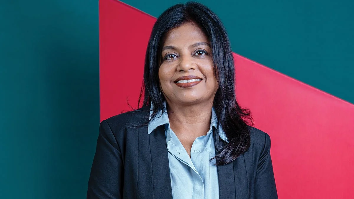 A woman with shoulder-length black hair smiling, dressed in a light blue collared shirt and a black blazer, standing in front of a teal and pink geometric background.