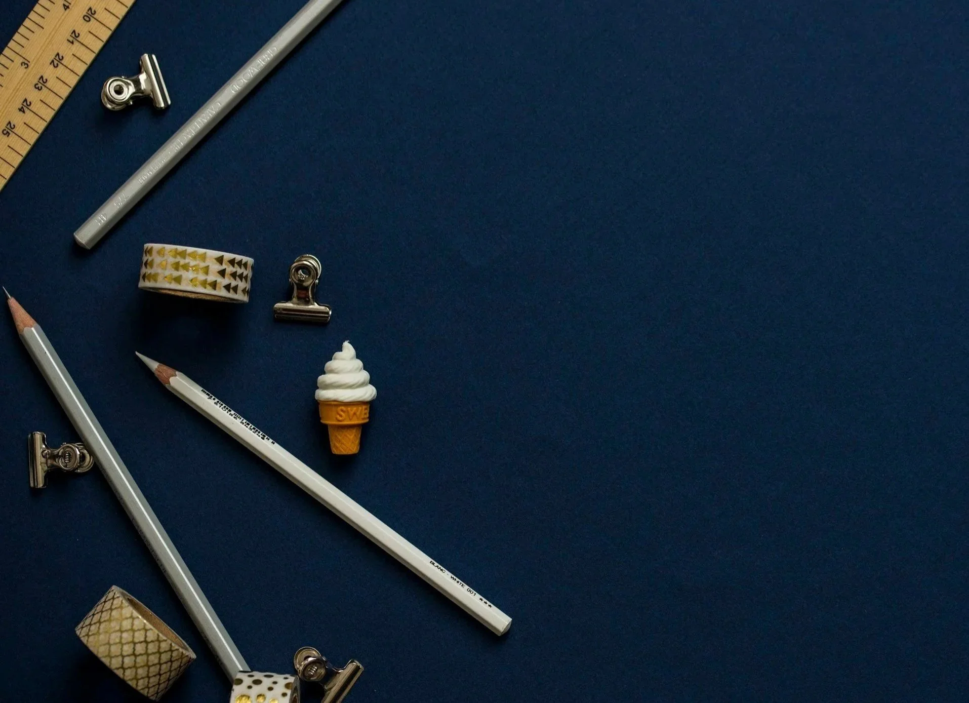 Flat lay of stationery items on a dark blue background, including pencils, paper clips, washi tapes, a ruler, and an ice cream-shaped eraser.